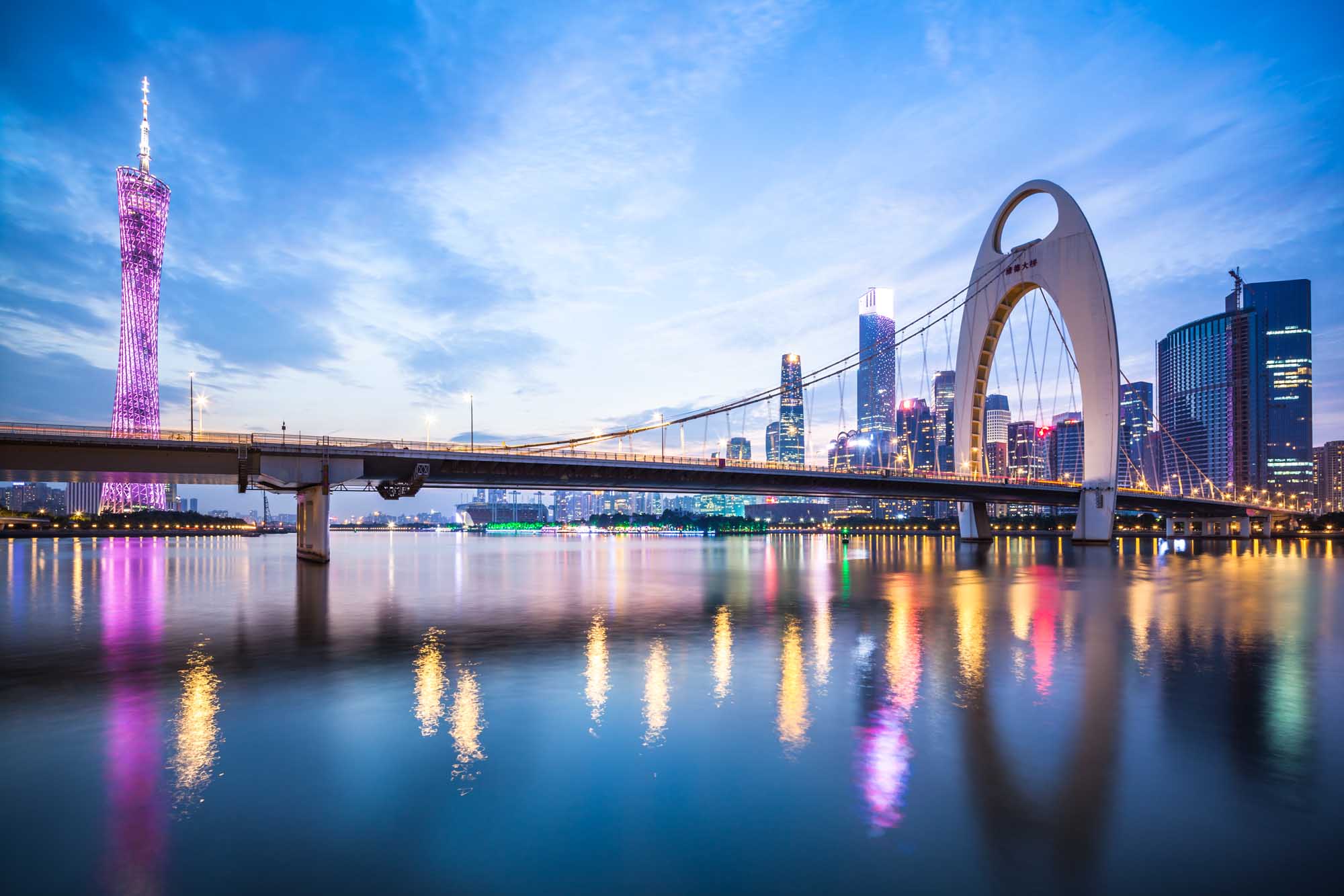 A breathtaking twilight cityscape of Guangzhou, China, featuring the iconic Liede Bridge spanning across the Pearl River. The scene captures the city's modern skyline illuminated with golden and pink lights, with a distinctive pink-lit tower on the left and a prominent white arch bridge on the right, all beautifully reflected in the calm waters below.