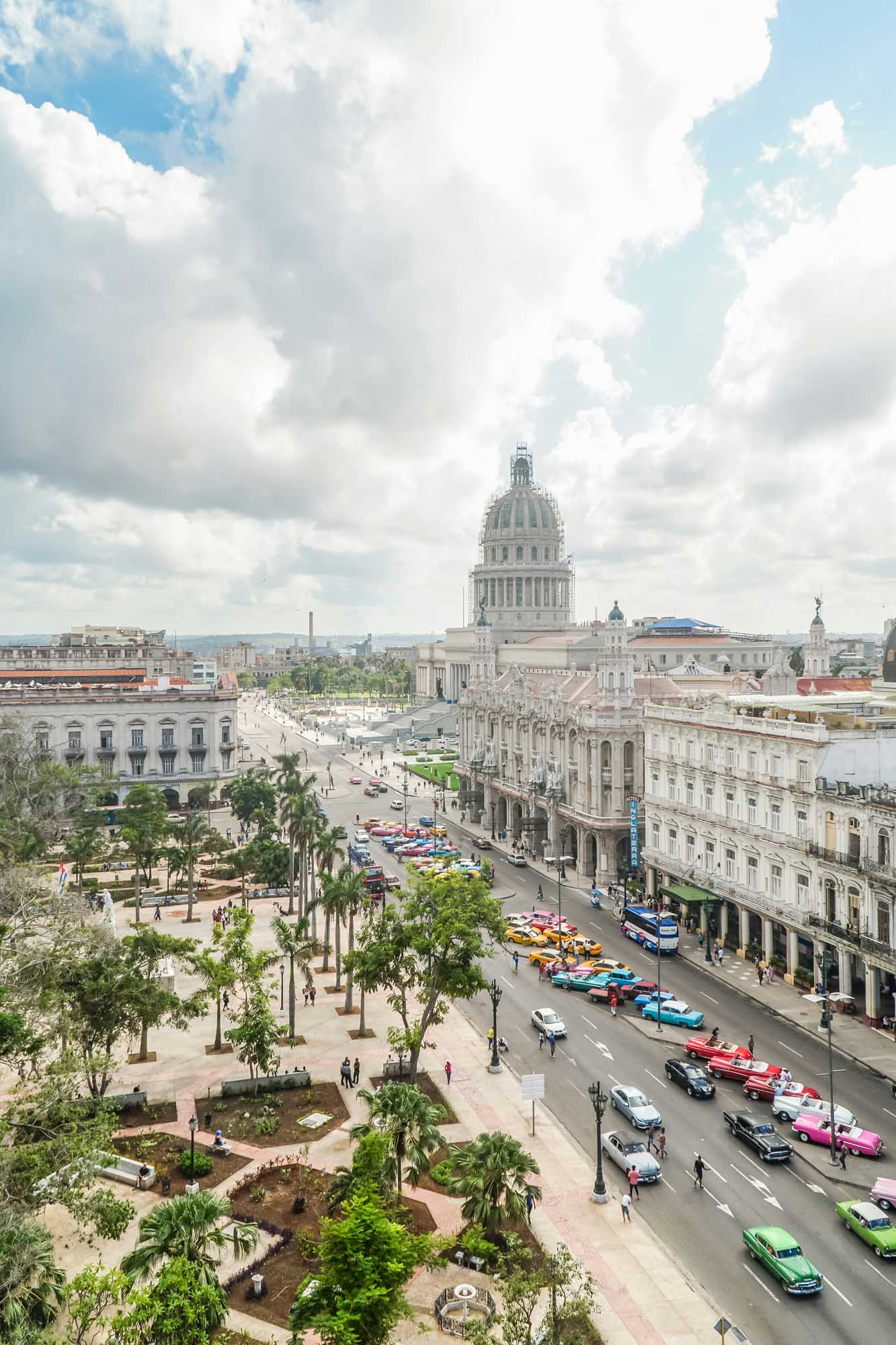 Havana Capitol Building with Classic American Cars An elevated daytime view of Havana's iconic Capitol building dominating the skyline, with its distinctive dome prominently featured. The street below bustles with colorful classic American cars and palm-lined plazas, capturing the vibrant energy of Cuba's capital city.