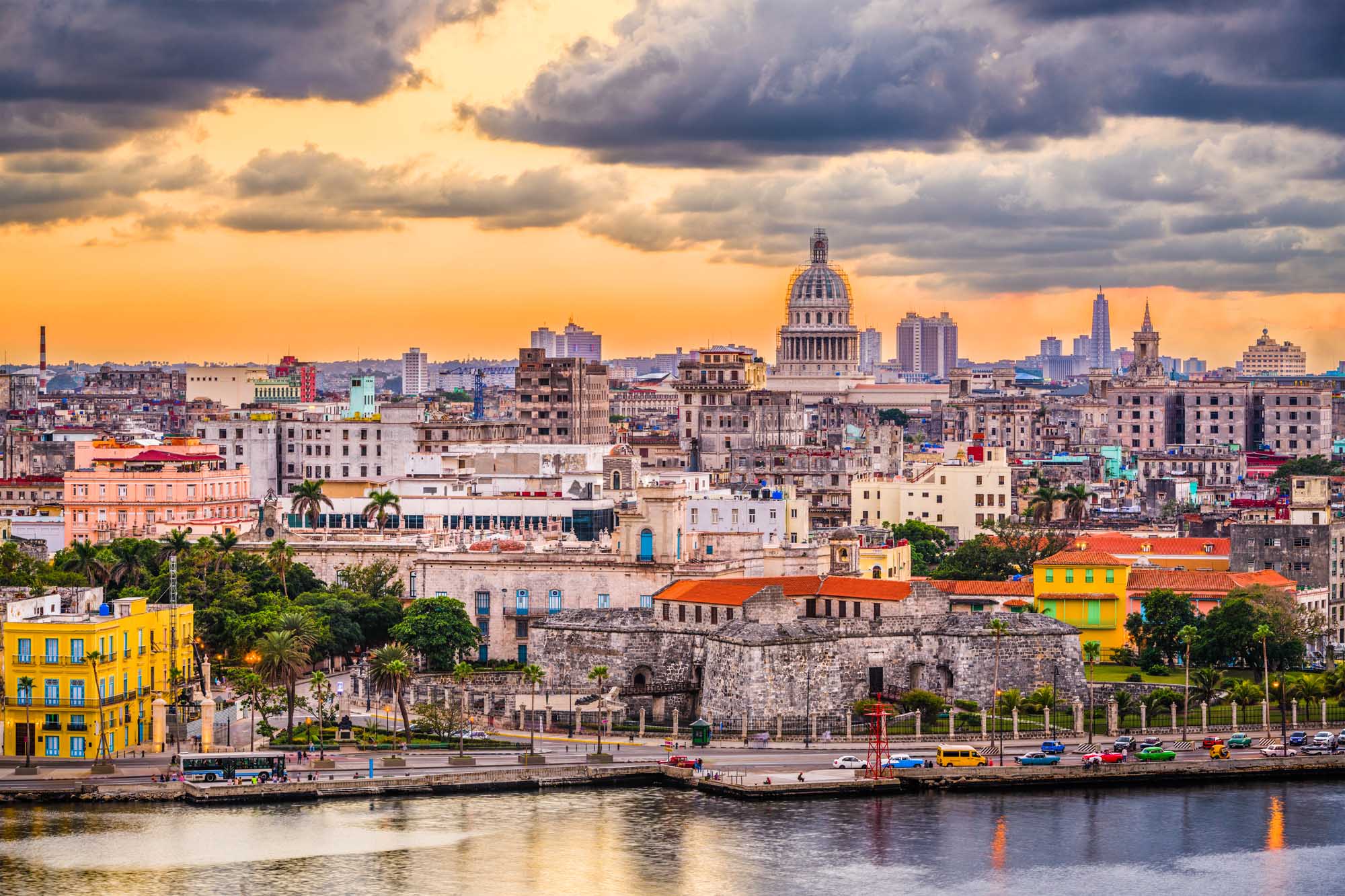 Havana Skyline at Sunset with Capitol Dome A stunning panoramic view of Havana, Cuba's downtown skyline at dusk, featuring the iconic Capitol building with its distinctive dome, colorful colonial architecture, palm trees, and the waterfront promenade (Malecón) along the harbor. The dramatic golden sky with storm clouds creates a striking backdrop for this vibrant Caribbean city.