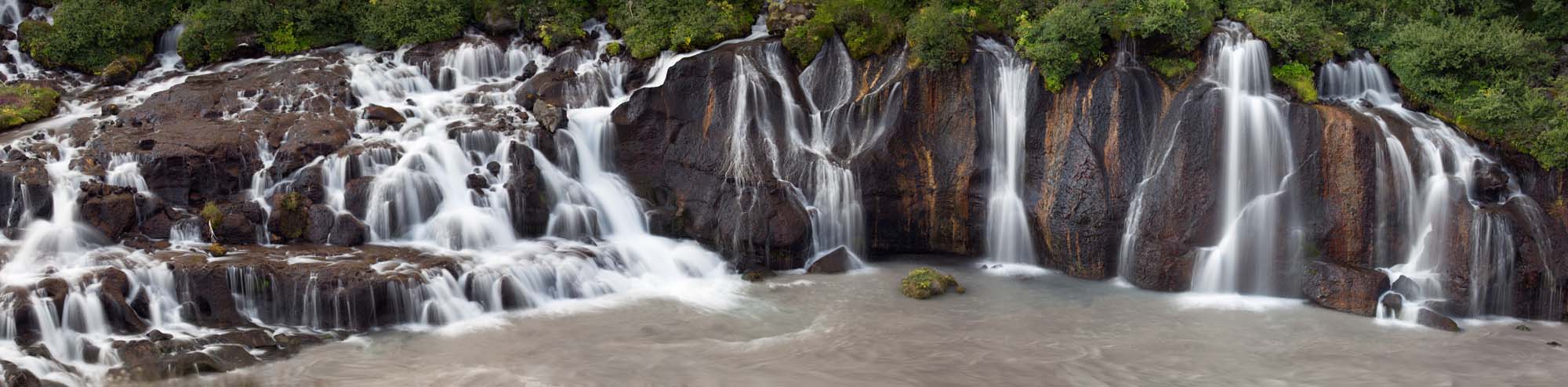 Hraunfossar Waterfall in Western Iceland A stunning cascade of multiple waterfalls flowing over dark basalt rock formations into a milky glacial river pool, surrounded by lush green vegetation. The image captures the dramatic natural beauty of Iceland's Hraunfossar waterfall system with its distinctive tiered drops and powerful water flow.