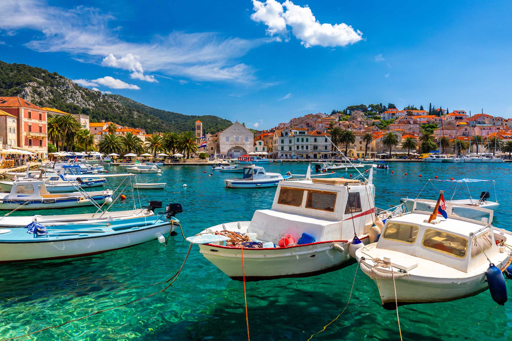 Hvar Harbor with Fishing Boats and Historic Town A picturesque Mediterranean harbor scene featuring numerous fishing boats anchored in crystal-clear turquoise waters off the coast of Hvar, Croatia. The historic town rises dramatically behind the harbor with traditional orange-roofed buildings nestled against a verdant hillside, showcasing the charming architecture and natural beauty of this popular Adriatic island destination.
