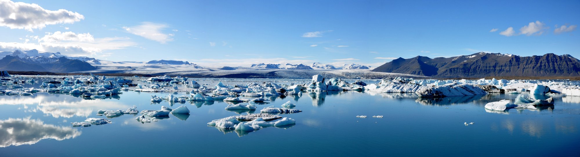 Jökulsárlón Glacier Lagoon Iceland Icebergs A breathtaking panoramic view of Jökulsárlón Glacier Lagoon in Iceland, featuring countless blue and white icebergs floating in pristine turquoise waters. Snow-capped mountains frame the horizon under a clear blue sky with scattered white clouds, showcasing one of Iceland's most stunning natural wonders.