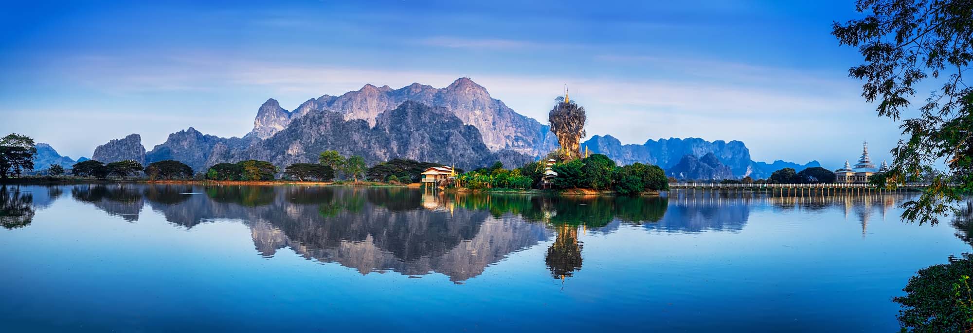 A stunning panoramic view of the iconic Kyauk Kalap Pagoda perched atop a dramatic limestone rock formation in Hpa-An, Myanmar, perfectly mirrored in the calm waters below. The golden pagoda and surrounding karst mountains create a serene and picturesque landscape under clear blue skies.