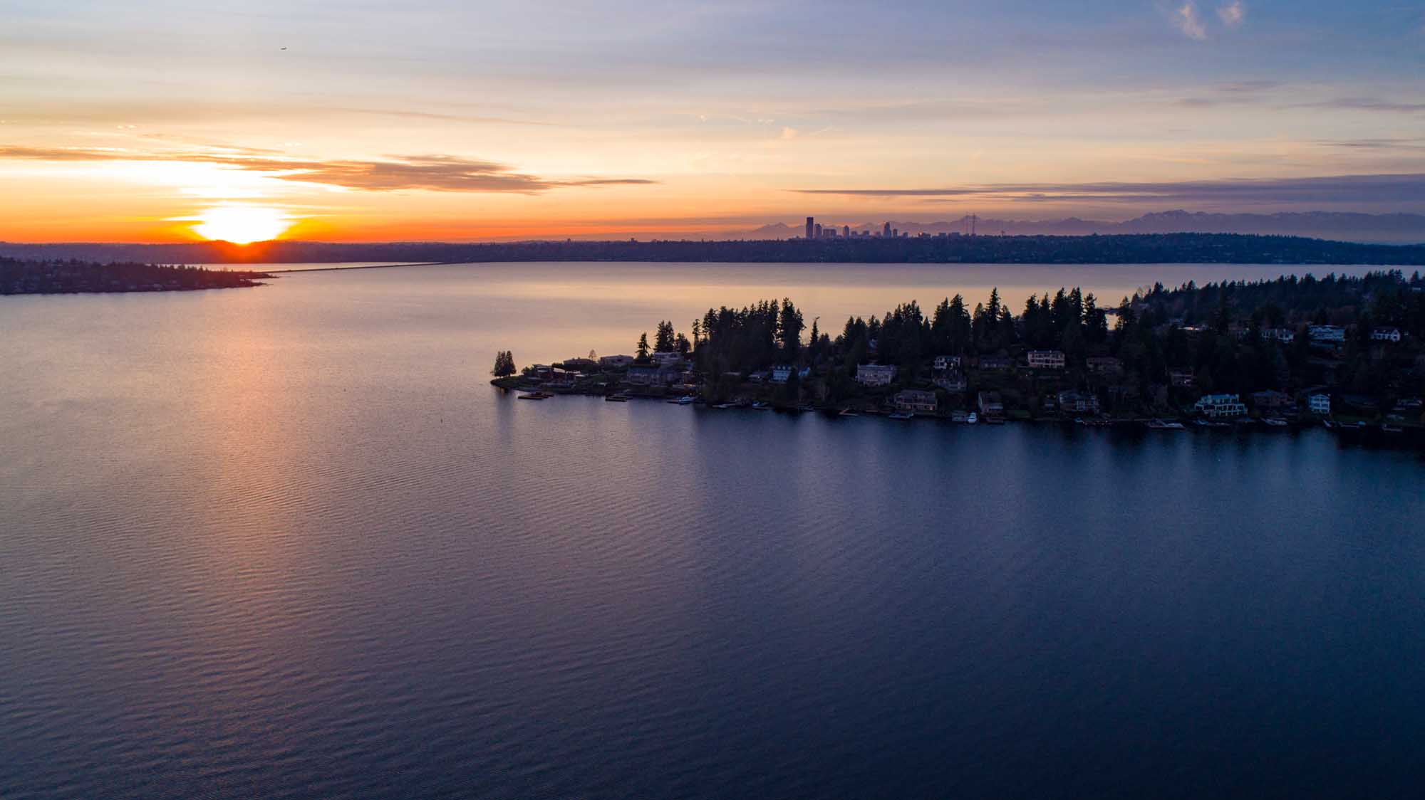 Lake Washington Sunset with Seattle Skyline A breathtaking sunset view over Lake Washington, featuring a residential waterfront community with dense evergreen forests in the foreground and the Seattle skyline silhouetted against a golden and purple sky across the water. This scenic vista captures the natural beauty of the Pacific Northwest with calm water reflections and the iconic Puget Sound region landscape.