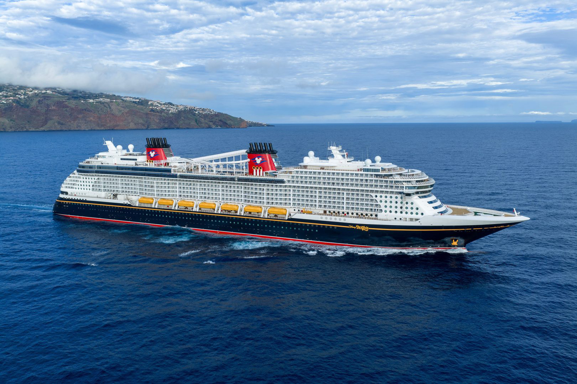 Disney Cruise Line Ship at Sea A massive modern cruise ship with distinctive red funnels featuring Mickey Mouse branding is anchored in brilliant blue waters off a volcanic island with lush settlements on its slopes. The vessel features a black hull with red striping, white superstructure, and yellow lifeboats, photographed under partly cloudy skies with a Mediterranean or Atlantic island landscape in the background.