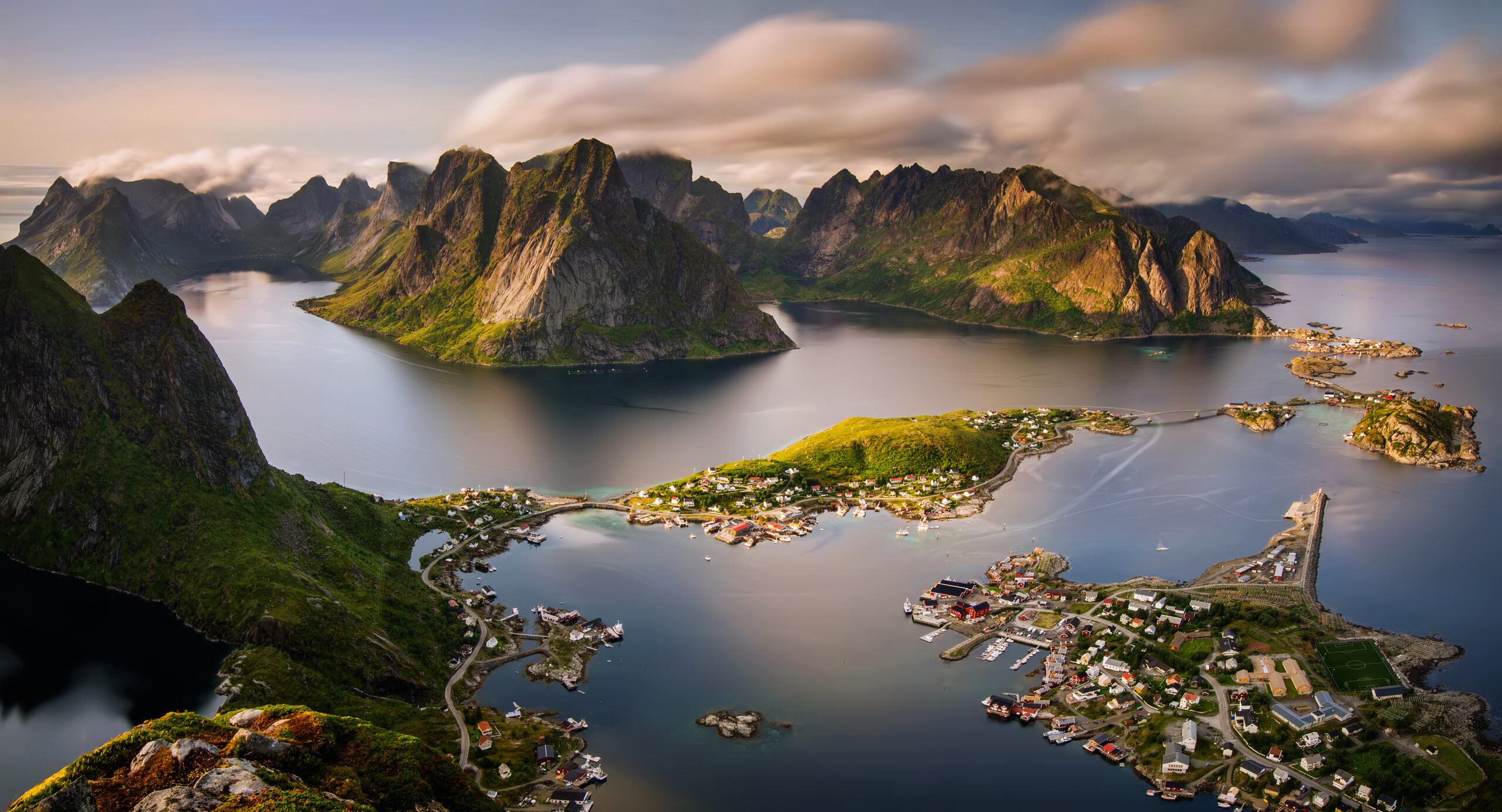 Lofoten Islands Aerial View with Coastal Villages A dramatic aerial photograph of the Lofoten Islands in northern Norway, showcasing towering jagged peaks rising from calm waters, with traditional fishing villages nestled along the coastline. The landscape features steep green-covered mountains, small red and yellow houses, and picturesque harbors surrounded by pristine Arctic waters.