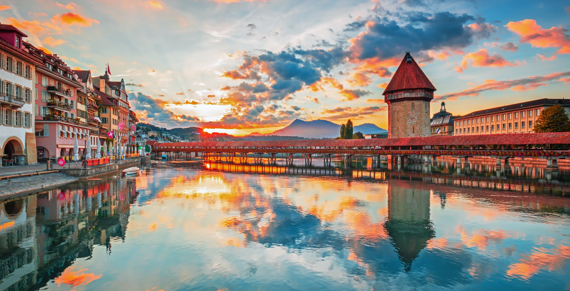 Lucerne Old Town Sunset with Chapel Bridge A stunning sunset view of Lucerne, Switzerland, featuring the iconic wooden Chapel Bridge (Kapellbrücke) with its distinctive red roof tower, colorful medieval buildings reflected in the calm waters of Lake Lucerne, and snow-capped mountains silhouetted against a vibrant golden and blue sky.