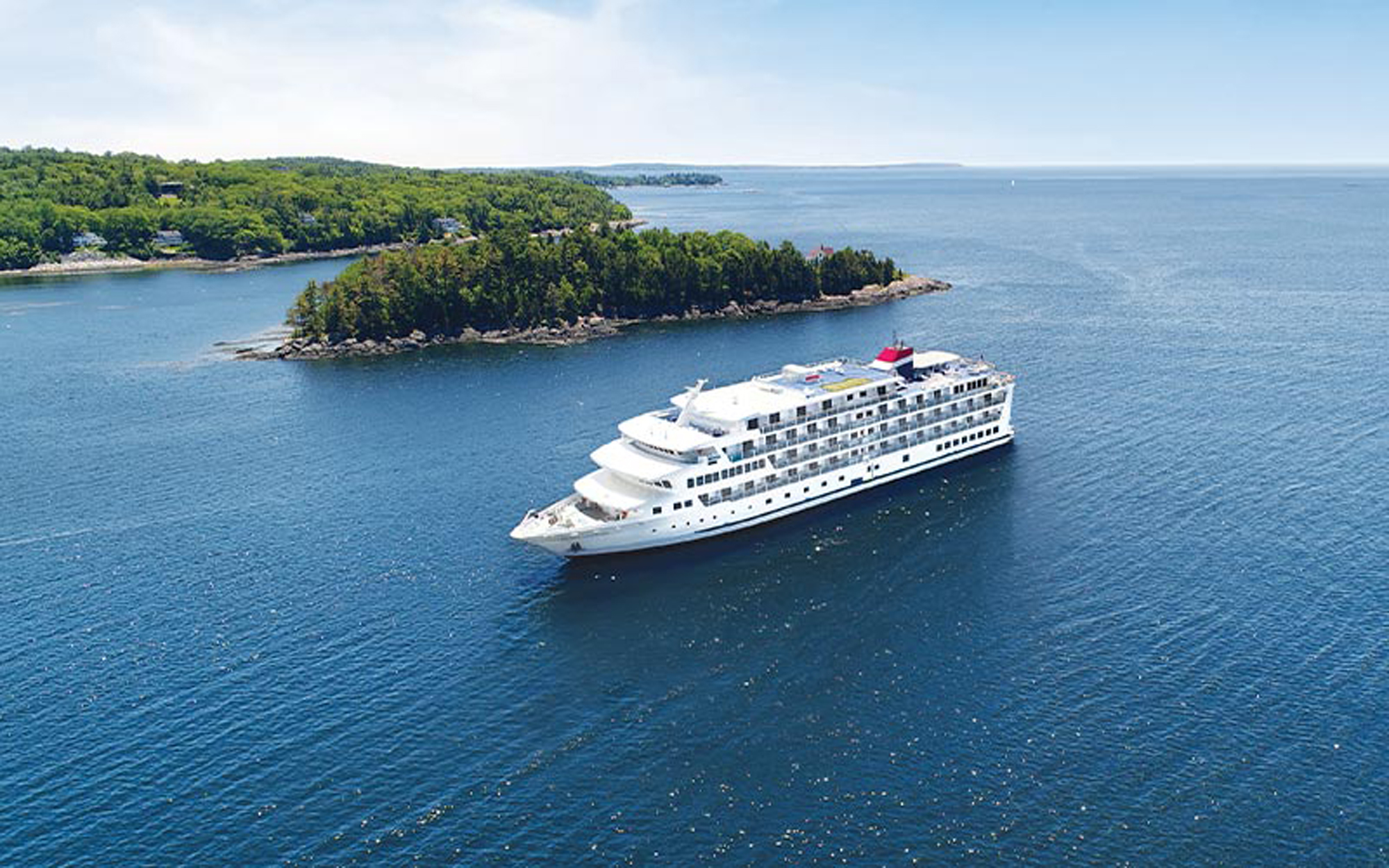 Luxury Cruise Ship in Puget Sound Waters A large, modern cruise ship with distinctive white hull and red funnel anchors in the calm blue waters of Puget Sound, with a forested peninsula dotted with residential homes visible in the background. The clear day and pristine waters showcase the natural beauty of the Pacific Northwest coastal region.