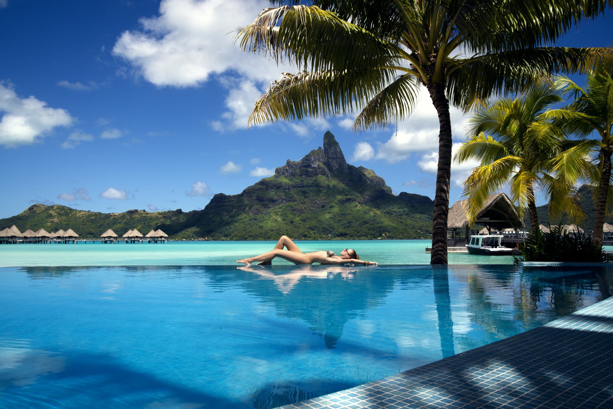 Luxury Resort Pool with Tropical Mountain View A woman relaxes on a floating lounger in an infinity pool at a tropical resort, with crystal-clear turquoise water, palm trees, and a dramatic volcanic peak in the background. The serene scene captures the essence of a luxurious South Pacific island getaway with overwater bungalows visible along the shoreline.