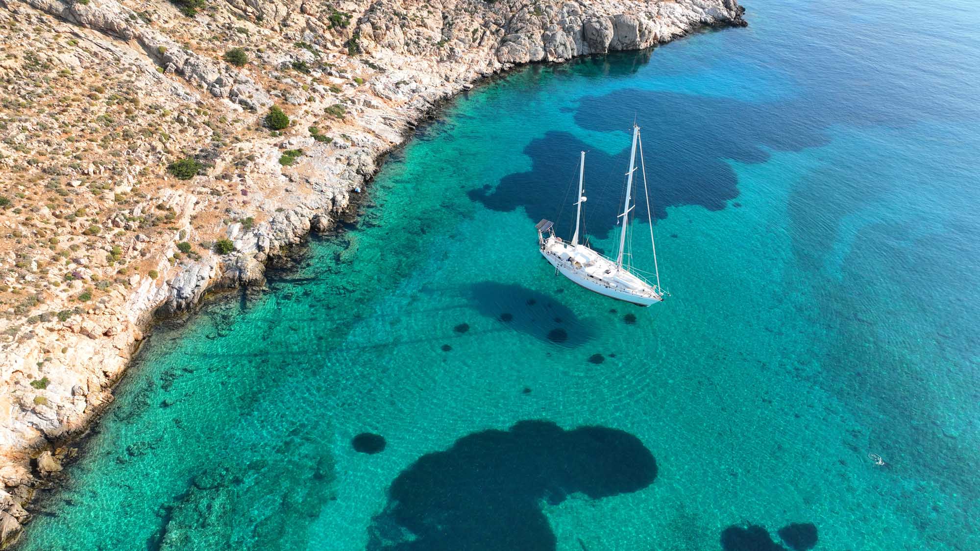 Luxury Sailboat Anchored in Mykonos Bay An aerial drone photograph of a white sailing yacht anchored in crystal-clear turquoise waters beside dramatic white limestone cliffs in Ornos Bay, Mykonos. The pristine Mediterranean seascape showcases the boat's reflection in the transparent water, with dark rocky outcrops visible beneath the surface.