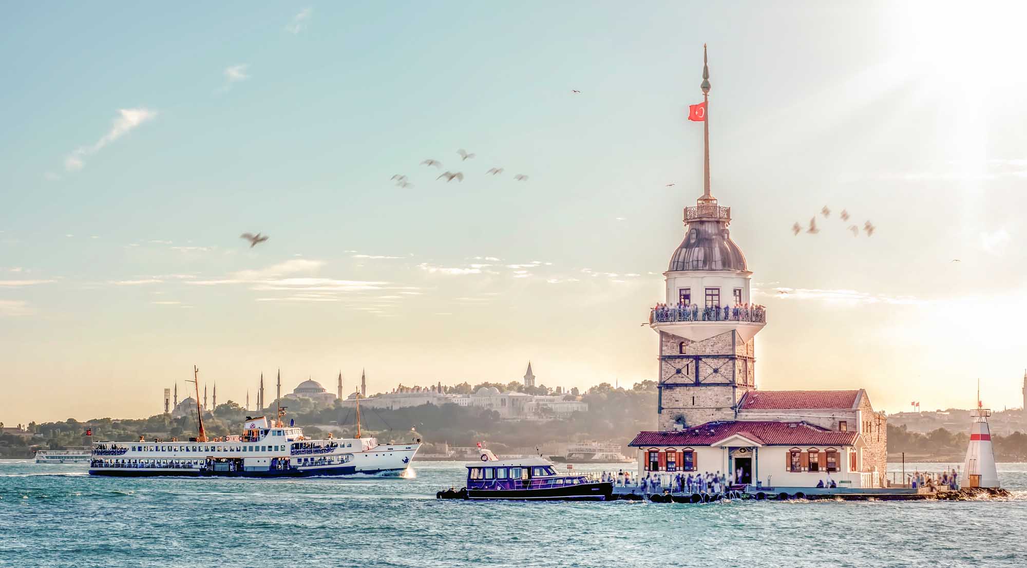 Maiden’s Tower Istanbul with Ferry Boats A scenic view of Istanbul's iconic Maiden's Tower (Kız Kulesi) in the Bosphorus Strait, with a blue and white passenger ferry approaching the tower and the cityscape of Istanbul visible in the background under a clear sky. The historic lighthouse-style tower features a domed top with Turkish flag, surrounded by water taxis and sightseeing vessels.