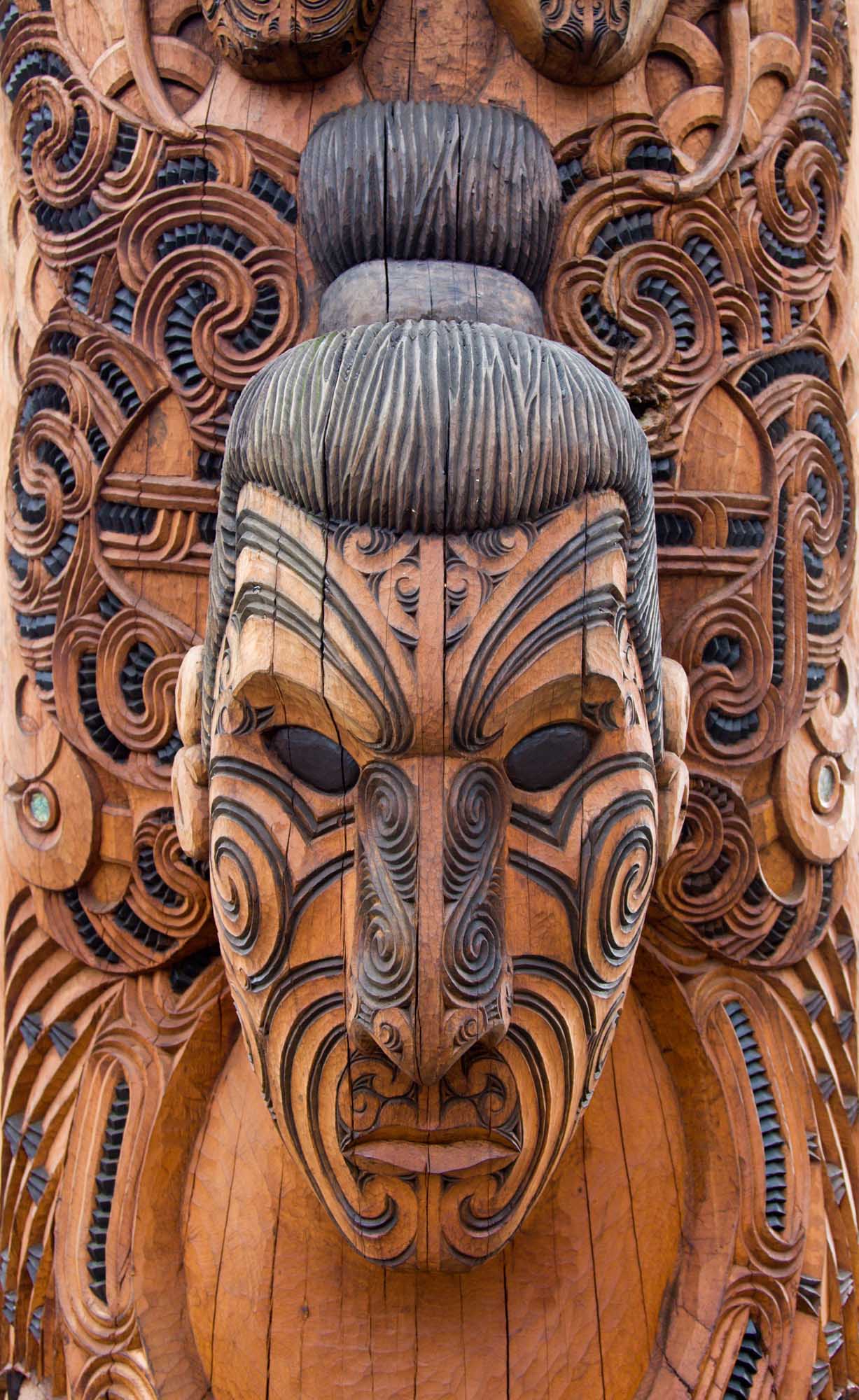 Maori Carved Wooden Face at Rotorua An intricately carved Maori wooden face featuring traditional geometric patterns, spiral designs, and a prominent top knot hairstyle. The carving displays characteristic Maori artistic motifs with detailed facial markings and elaborate decorative elements in warm wood tones.