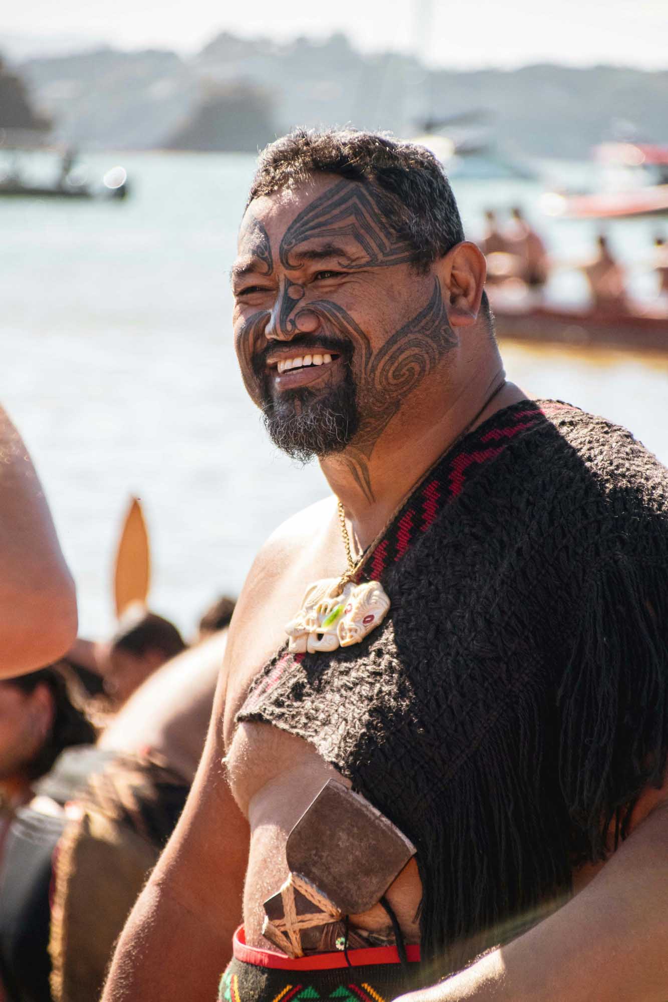 Maori Man with Traditional Facial Tattoo A man with traditional Maori ta moko facial tattoos smiles at the camera while standing near a waterfront with boats and hills in the background. He wears traditional Maori garments including a woven cloak and ornamental pendant, representing indigenous New Zealand culture.