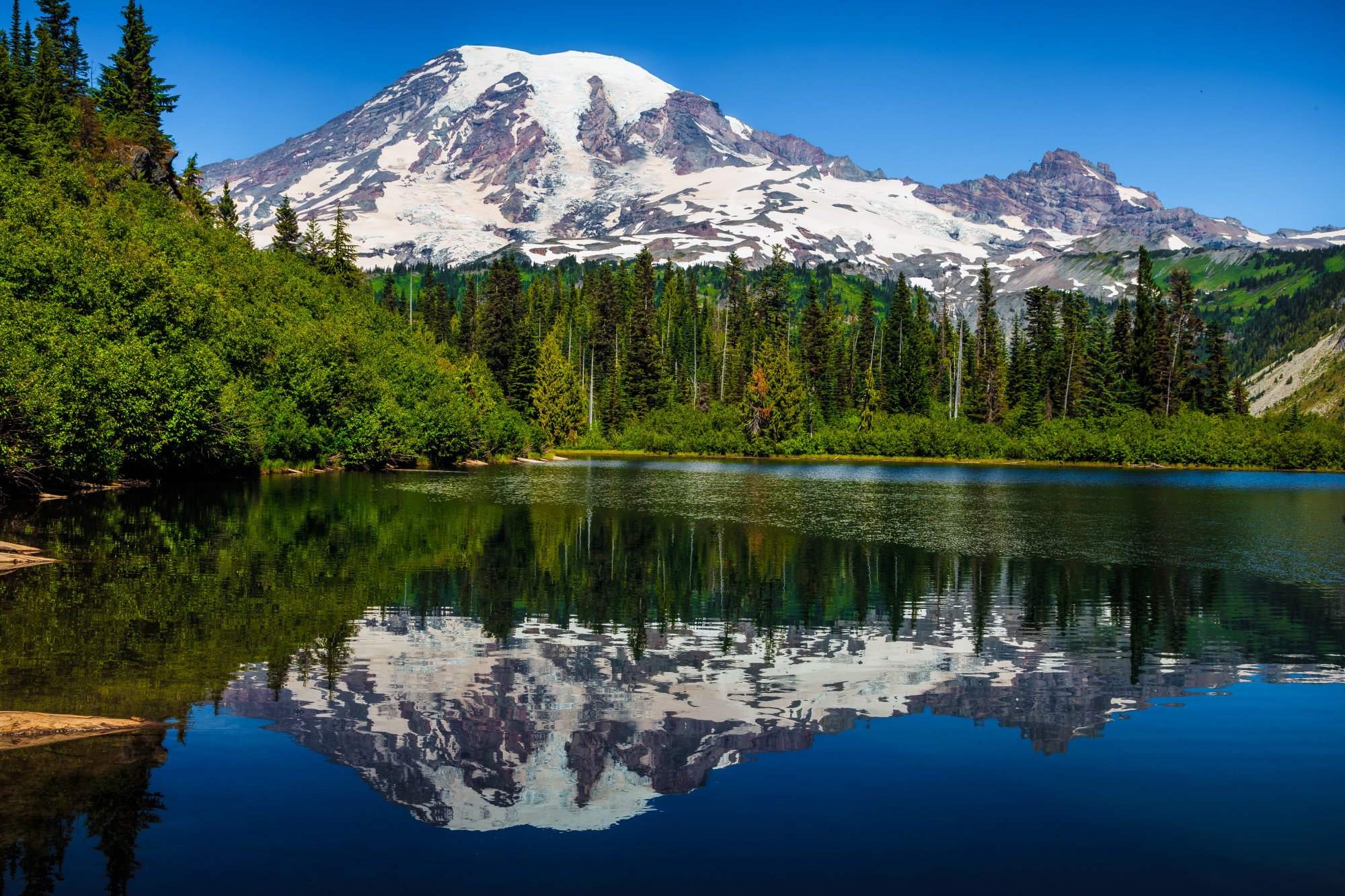 Mount Rainier Reflection in Alpine Lake A stunning alpine landscape featuring Mount Rainier's snow-capped summit perfectly reflected in a pristine mountain lake, surrounded by dense coniferous forest under clear blue skies. The scene captures the dramatic contrast between the glaciated peak, evergreen forests, and mirror-like water.