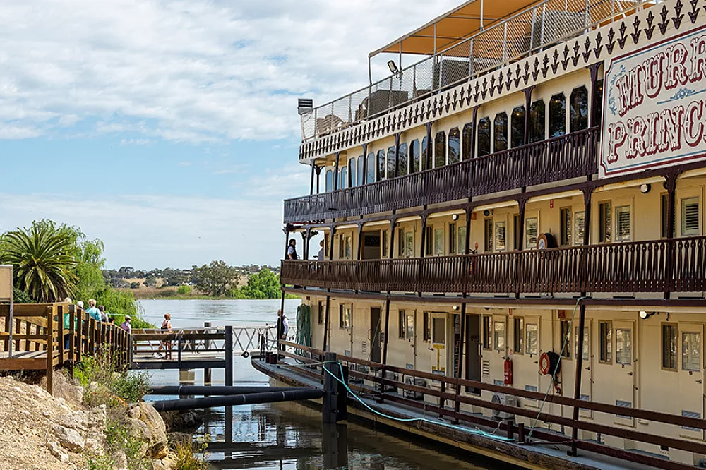 Murray Princess Riverboat Docked at Riverbank A three-deck paddlewheel riverboat with cream and brown colonial-style architecture docked along a peaceful river, featuring ornate railings and multiple levels of verandahs. The vessel displays classic Australian riverboat design with traditional paddle-wheel aesthetics and serves as a floating accommodation for river cruises.