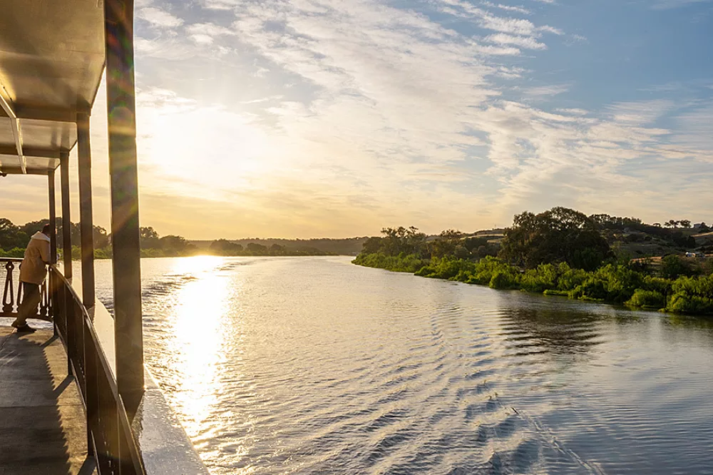 Murray River Sunset Views from Deck A scenic view of the Murray River at golden hour, captured from the deck of a riverboat. The sun reflects beautifully across the calm water, with lush green vegetation lining both riverbanks under a partly cloudy sky.