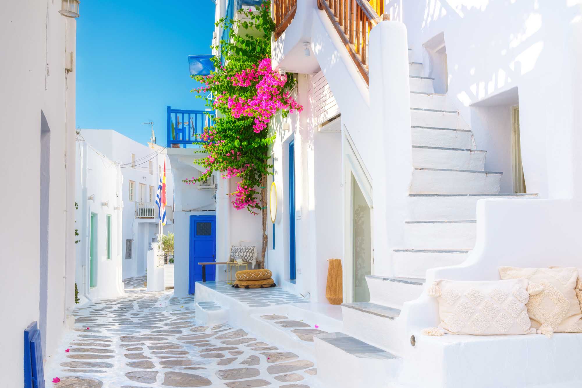 Mykonos Whitewashed Street with Bougainvillea A charming narrow alleyway in Mykonos, Greece, featuring quintessential Cycladic architecture with whitewashed buildings, blue doors and shutters, and vibrant magenta bougainvillea cascading down a building. The traditional cobblestone street, bright Mediterranean sunlight, and picturesque Greek island ambiance make this an iconic travel destination scene.