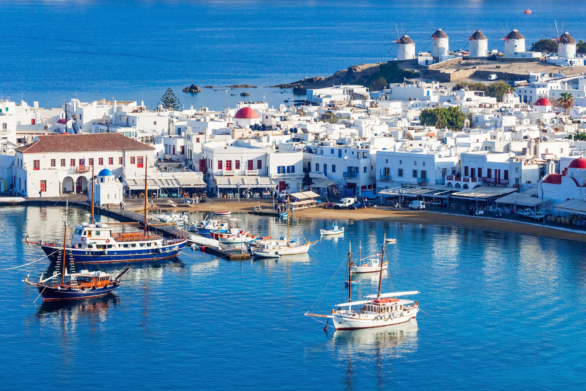 Mykonos Windmills and Harbor Vista A picturesque view of Mykonos, Greece, featuring the iconic whitewashed windmills perched on a hillside overlooking the turquoise harbor filled with traditional fishing boats and sailboats. The charming Cycladic architecture with blue-domed roofs and white-washed buildings cascades down to the waterfront, creating a quintessential Mediterranean island scene.