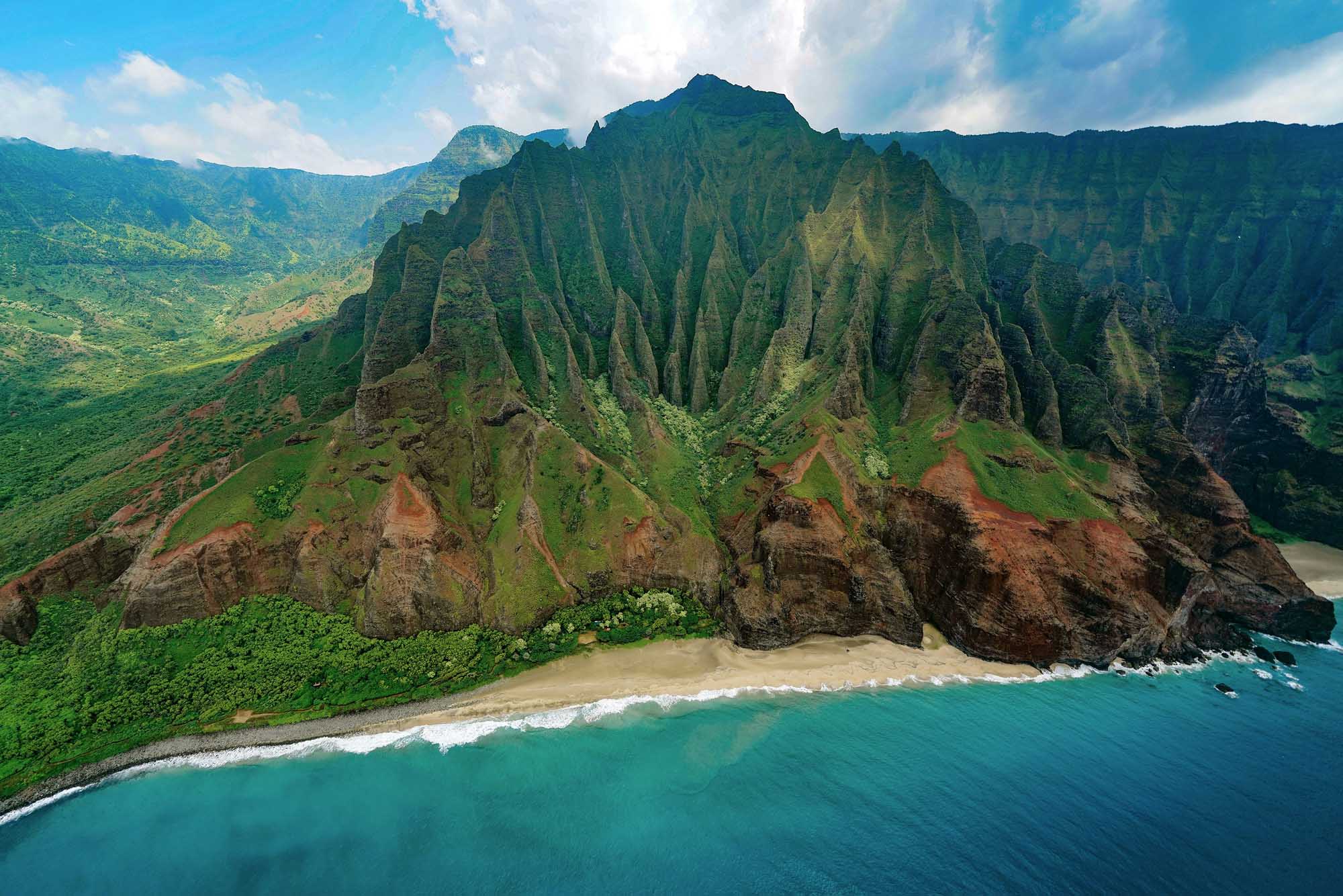 Na Pali Coast Cliffs and Turquoise Waters A stunning aerial view of the iconic Na Pali Coast featuring dramatic emerald-green fluted sea cliffs rising vertically from a pristine sandy beach and turquoise Pacific Ocean waters. The distinctive sharp ridges and valleys carved into the mountainous terrain create a striking natural landscape characteristic of this UNESCO World Heritage site.