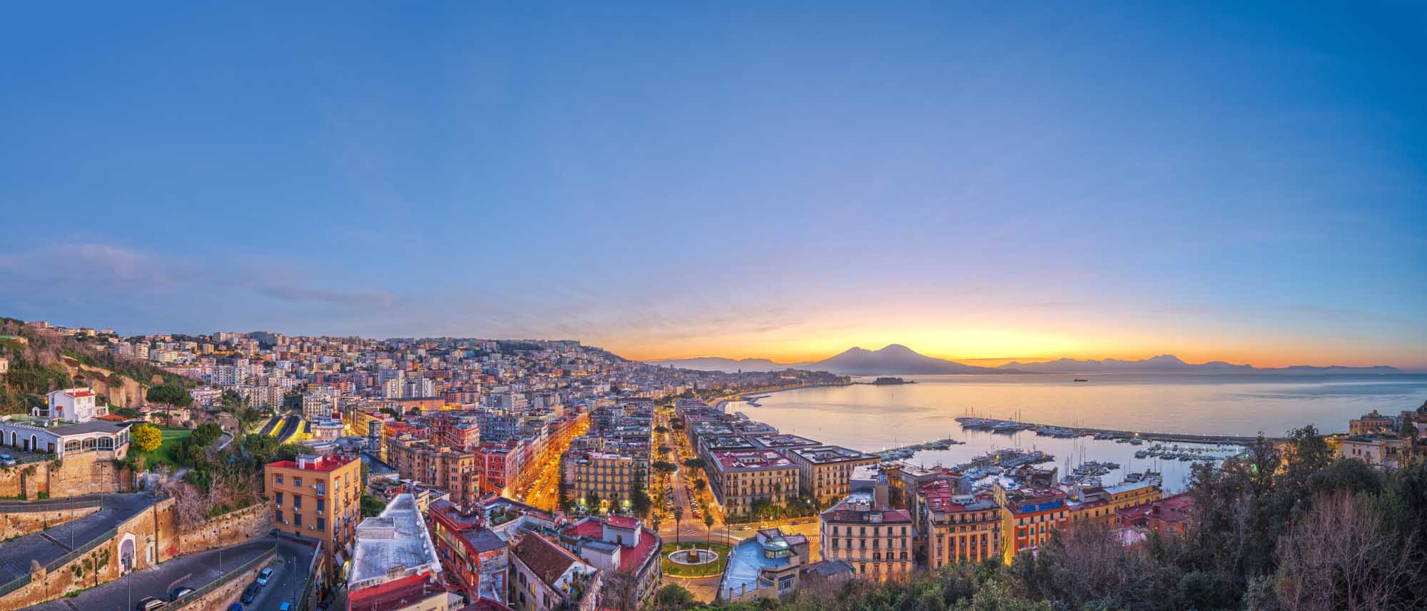 Naples Skyline at Dawn with Mount Vesuvius An expansive aerial view of Naples, Italy at sunrise, showcasing the densely-packed colorful buildings cascading down hillsides toward the Bay of Naples. Mount Vesuvius is visible on the horizon across the bay, silhouetted against the golden dawn sky, while numerous sailboats dot the harbor below.