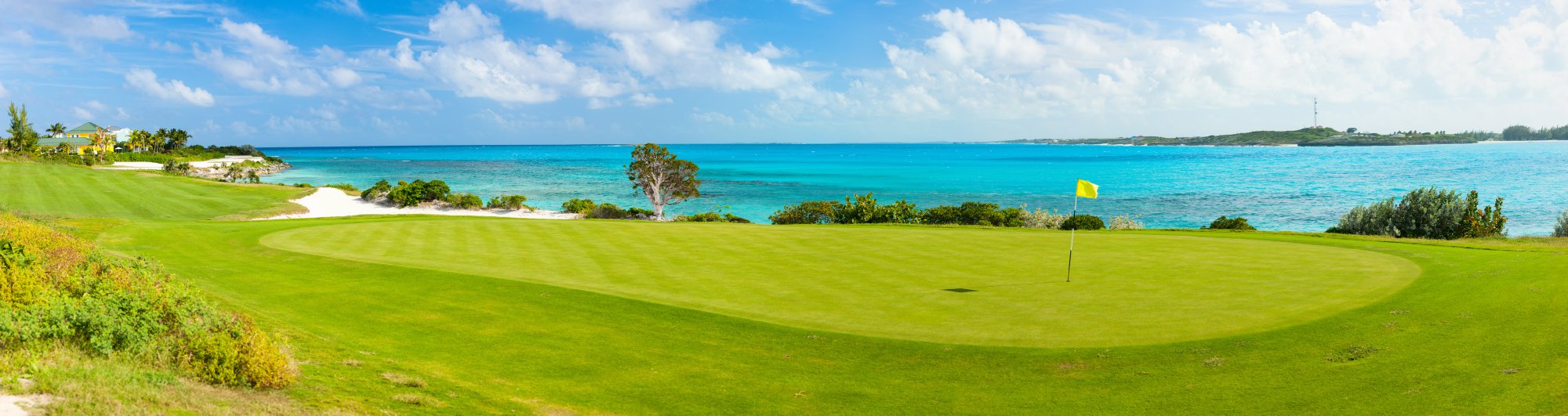 Oceanfront Golf Course with Turquoise Waters A scenic coastal golf course featuring a pristine green fairway overlooking crystal-clear turquoise waters and a white sand beach, with lush tropical vegetation and a yellow flag marking the pin in the distance.