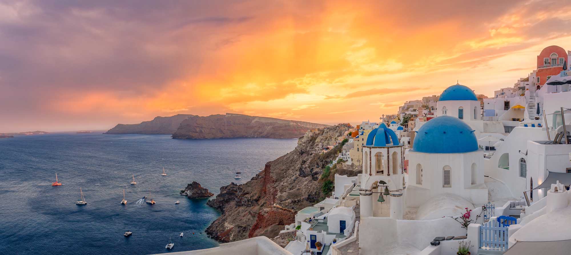 Oia Sunset Over Santorini Caldera A breathtaking panoramic view of the iconic village of Oia perched on the cliffs of Santorini, Greece, featuring traditional whitewashed buildings with distinctive blue-domed churches overlooking the Aegean Sea during a stunning golden sunset. Sailboats dot the crystal-clear waters below, creating a quintessential Mediterranean luxury travel destination scene.