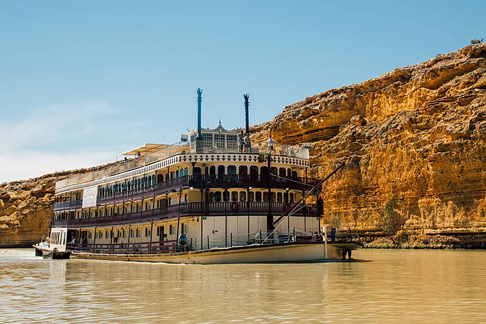 Murray Princess Riverboat on Golden Cliffs A classic multi-deck paddlewheel riverboat with distinctive twin smokestacks navigates a muddy river, with dramatic golden-hued limestone cliffs towering in the background. The vessel features traditional stern-wheel design with ornate white railings and multiple passenger decks typical of Mississippi River-style cruising.