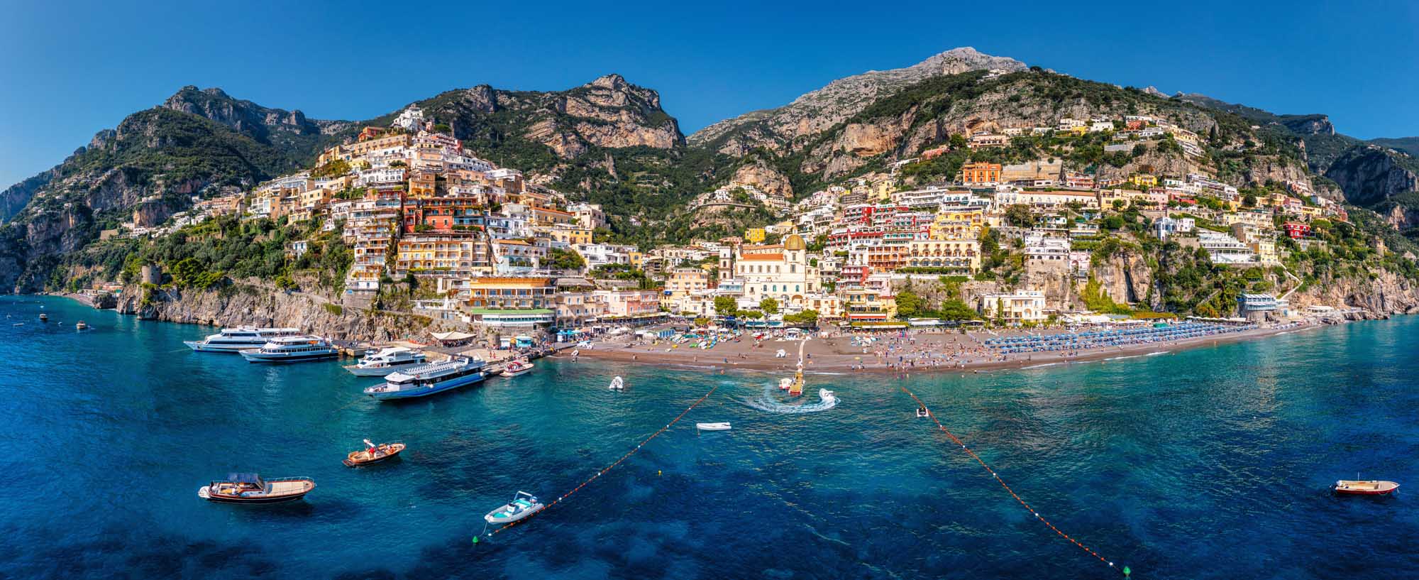 Positano Cliffside Village on Amalfi Coast An aerial panoramic view of Positano, a picturesque cliffside village perched dramatically on the Amalfi Coast in Italy. The colorful cascading buildings descend steeply toward a sandy beach and turquoise Mediterranean waters dotted with boats, showcasing the iconic architecture and stunning coastal landscape characteristic of Campania.