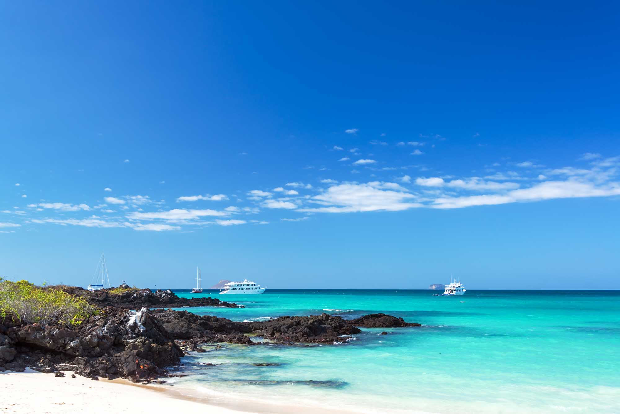 Pristine Beach with Anchored Boats in Galapagos A stunning tropical beach scene featuring crystal-clear turquoise waters, dark volcanic rock
