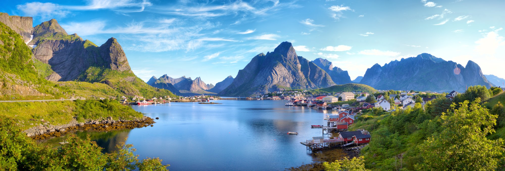 Reine Village in Lofoten Islands, Norway A stunning panoramic view of Reine, a picturesque fishing village nestled in the Lofoten Islands of northern Norway, featuring traditional red cabins built over the water, dramatic jagged mountain peaks, and calm blue fjord waters under a clear sky.