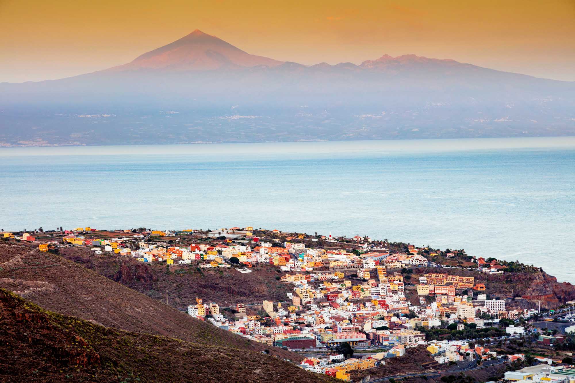 San Sebastián de la Gomera with Teide Volcano A picturesque coastal town cascades down volcanic slopes toward turquoise Atlantic waters, with its distinctive colorful buildings in vibrant reds, yellows, and whites. In the distance across the ocean, the majestic snow-capped peak of Mount Teide on Tenerife dominates the horizon under a golden sunrise sky.