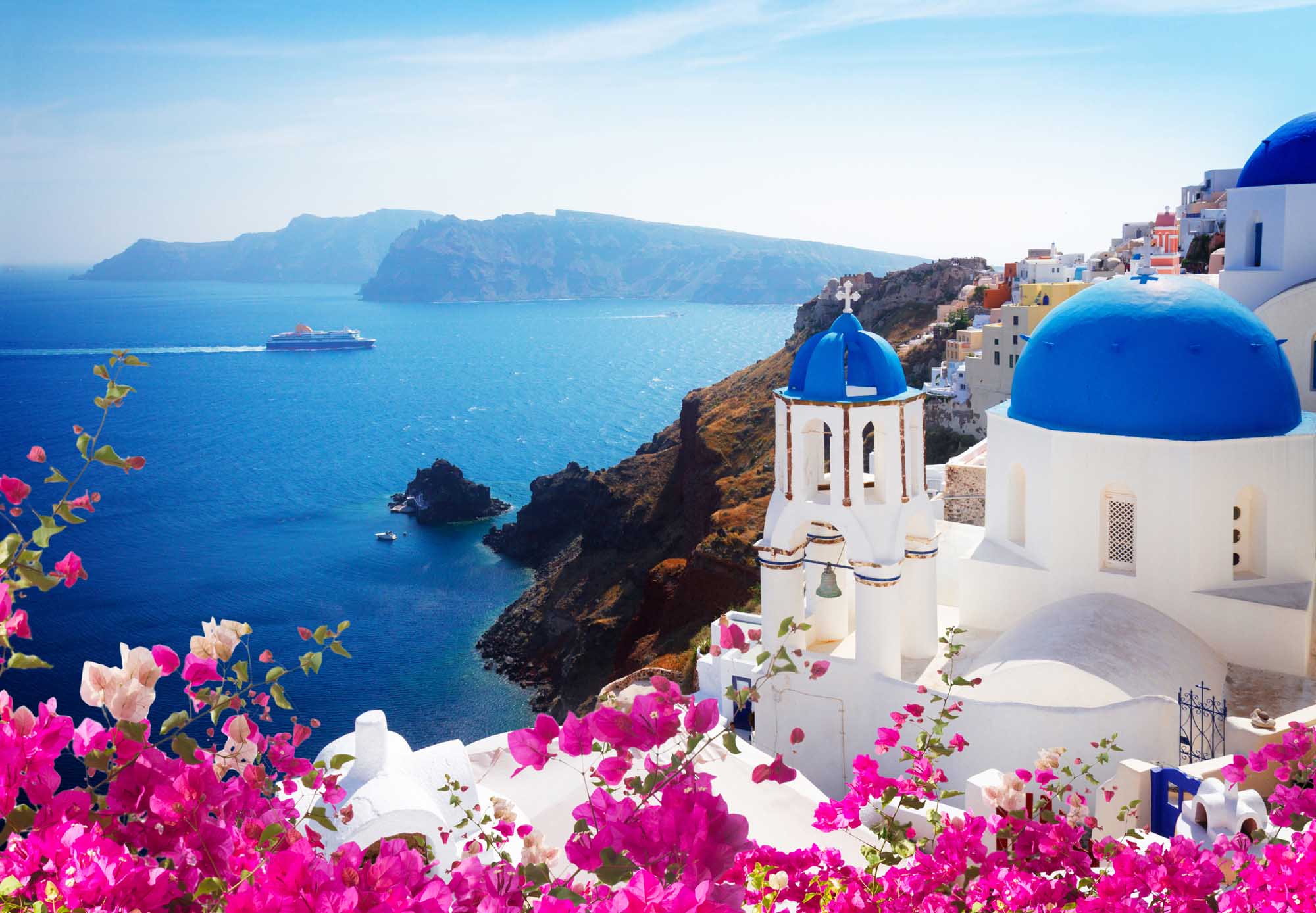 Santorini Cliffside Village with Blue-Domed Church A picturesque view of Oia village in Santorini, Greece, featuring iconic white-washed buildings with brilliant blue-domed churches overlooking the deep blue Aegean Sea. Vibrant magenta bougainvillea flowers frame the foreground, with a cruise ship visible in the distance and the volcanic caldera landscape providing a dramatic backdrop.