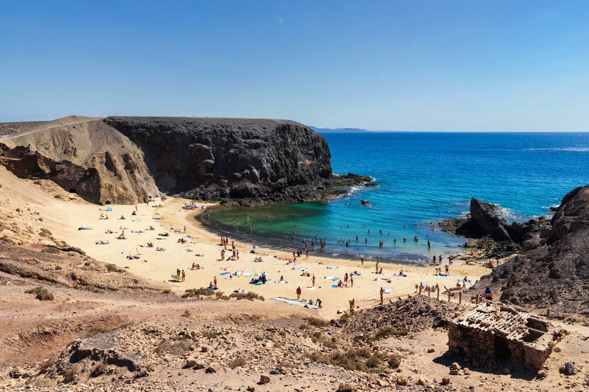 Secluded Cove Beach with Dramatic Cliffs A pristine sandy beach nestled beneath towering dark volcanic cliffs, featuring crystal-clear turquoise waters and numerous sunbathers enjoying the Mediterranean summer. The sheltered cove is surrounded by rugged rock formations, creating a dramatic and picturesque coastal landscape.