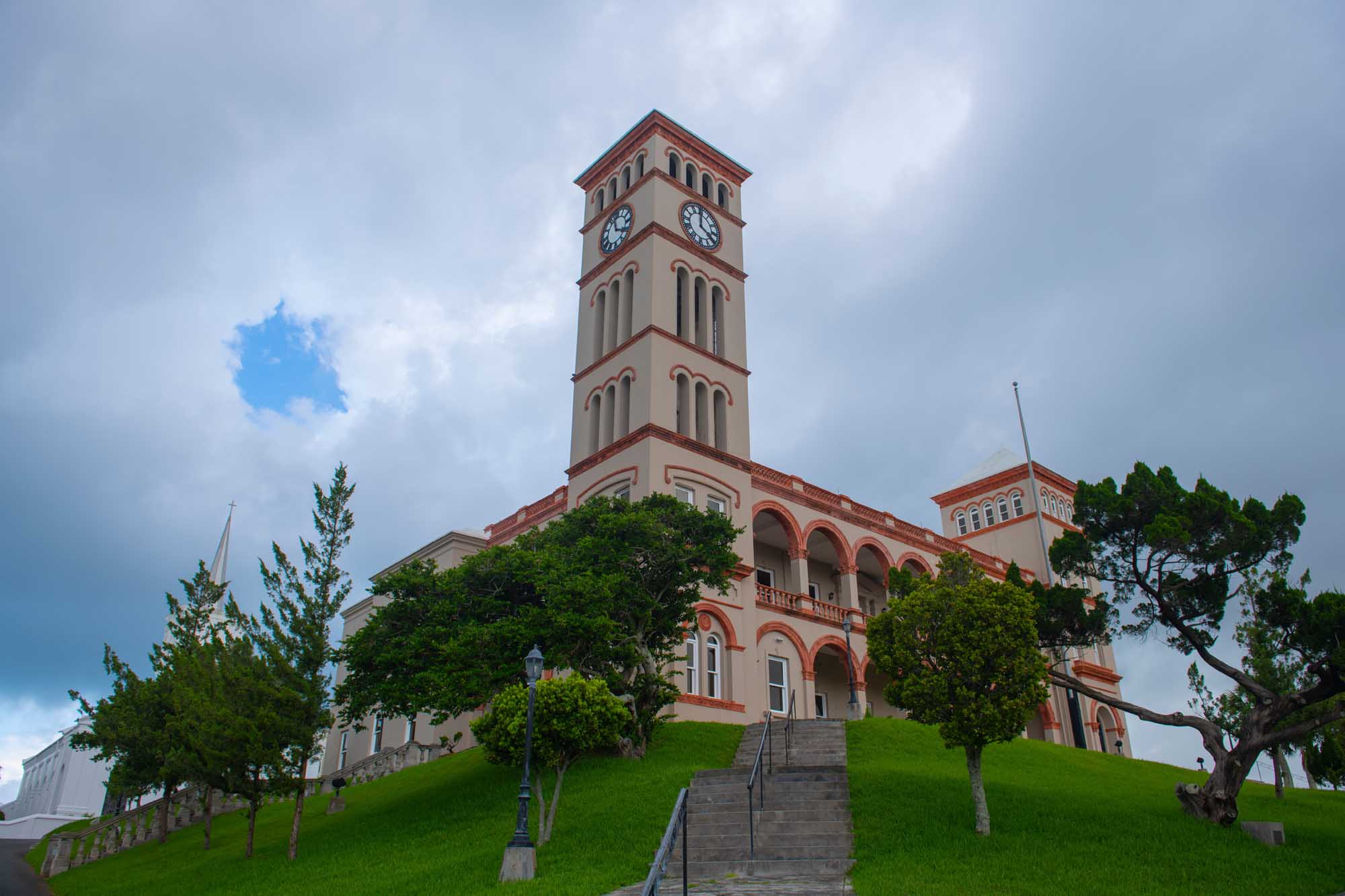 Sessions House Clock Tower in Hamilton, Bermuda A distinctive pink and cream-colored colonial government building featuring a prominent clock tower, situated on a manicured green hillside in Hamilton's city center. The Sessions House serves as the home of the House of Assembly and Supreme Court of Bermuda, showcasing classic Bermudian architecture with arched windows and ornate details.