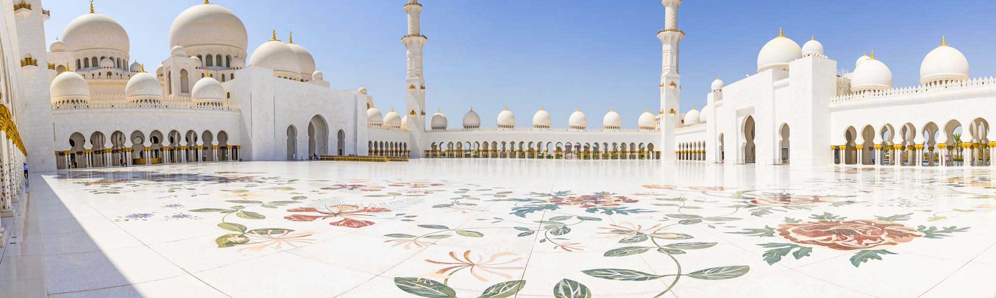 Sheikh Zayed Grand Mosque Courtyard The stunning courtyard of Sheikh Zayed Grand Mosque in Abu Dhabi, featuring pristine white marble flooring adorned with intricate floral inlay patterns in colorful stones. The mosque's iconic white domes, minarets, and ornate golden architectural details are visible under a clear blue sky, creating a serene and majestic Islamic architectural landscape.