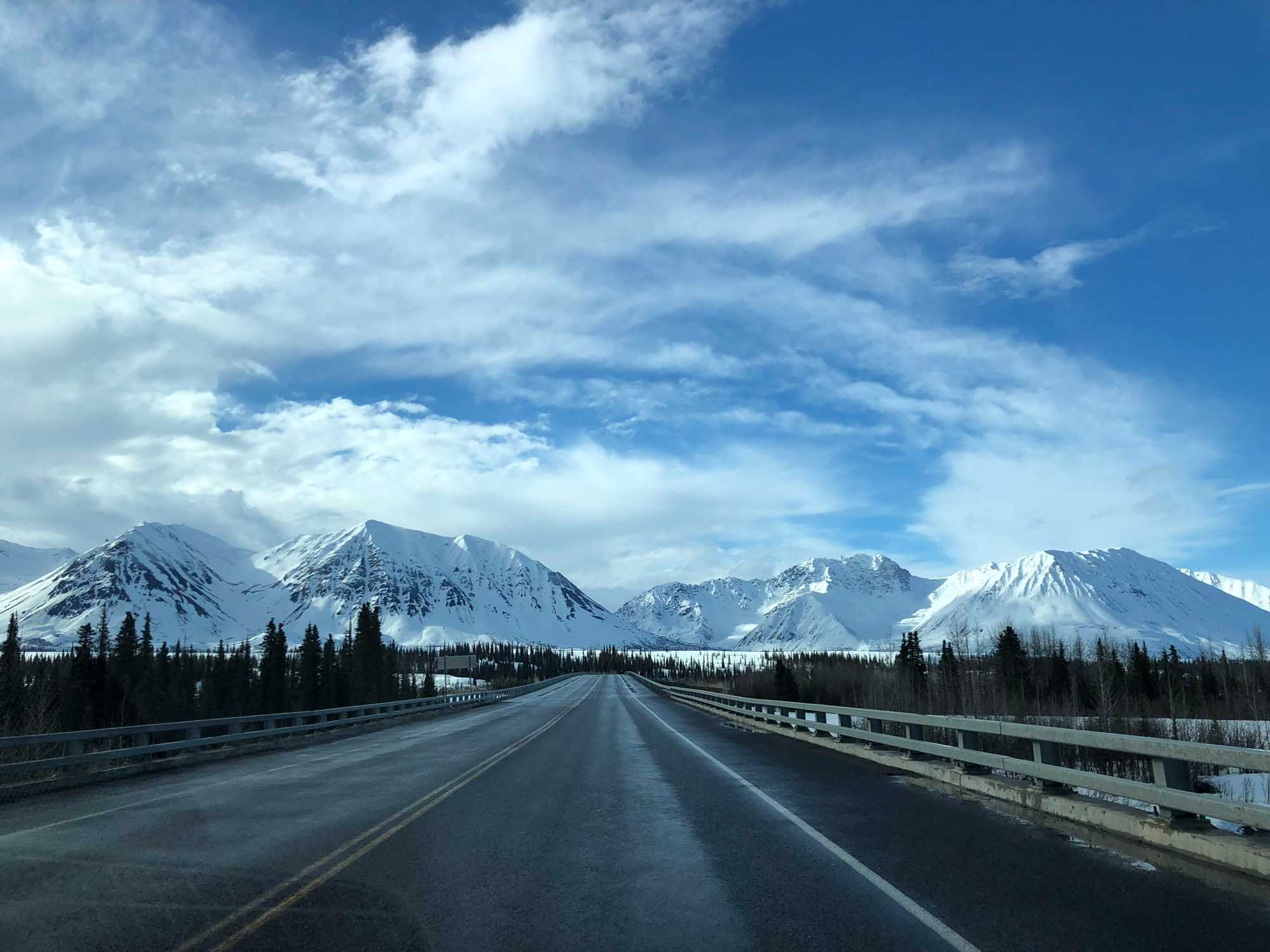 Snow-Capped Mountains Along Alaska Highway A straight highway stretches toward a dramatic range of snow-covered volcanic peaks under a brilliant blue sky with wispy clouds. Dense coniferous forests line both sides of the road, with metal guardrails visible along the bridge or elevated section, creating a striking perspective toward the distant mountains.