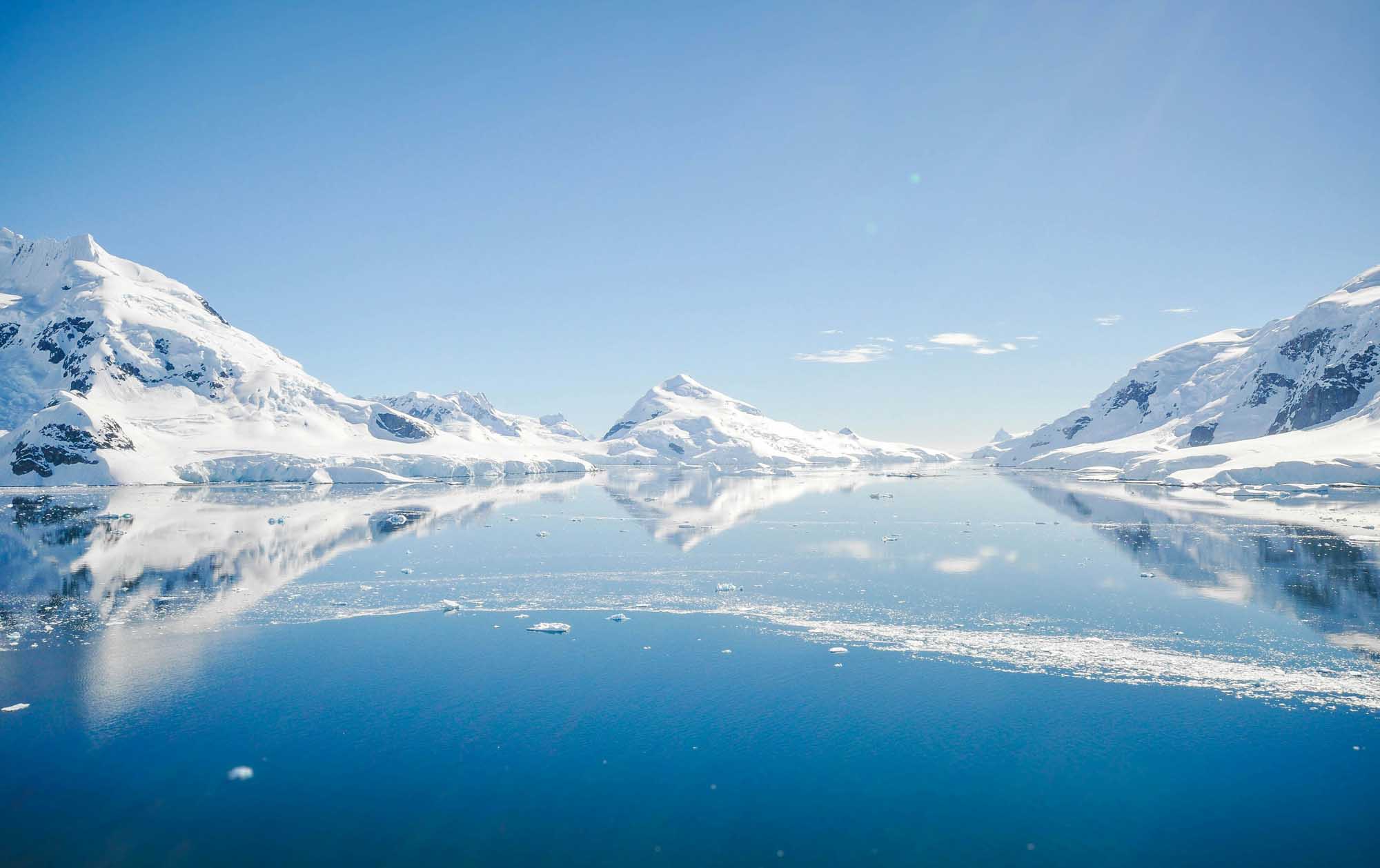 Snow-Capped Mountains and Icebergs in Antarctic Waters A breathtaking Arctic or Antarctic landscape featuring pristine snow-covered mountains reflected in calm, clear waters dotted with icebergs. The serene scene captures the majesty of polar regions with towering peaks and glacial ice under a bright, clear blue sky.