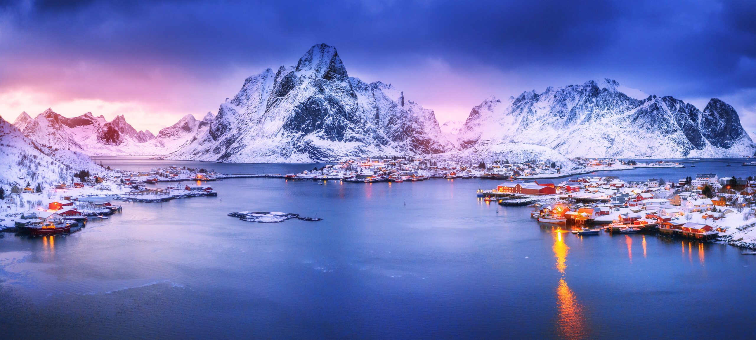 Snowy Mountains Over Arctic Fishing Village A stunning winter landscape featuring snow-capped mountains towering over a picturesque Arctic fishing village with traditional red and yellow wooden houses reflected in calm blue waters during twilight. The scene captures the dramatic beauty of Norway's Lofoten Islands with their characteristic peaks and traditional fishermen's cabins (rorbuer) illuminated by the soft glow of sunset.