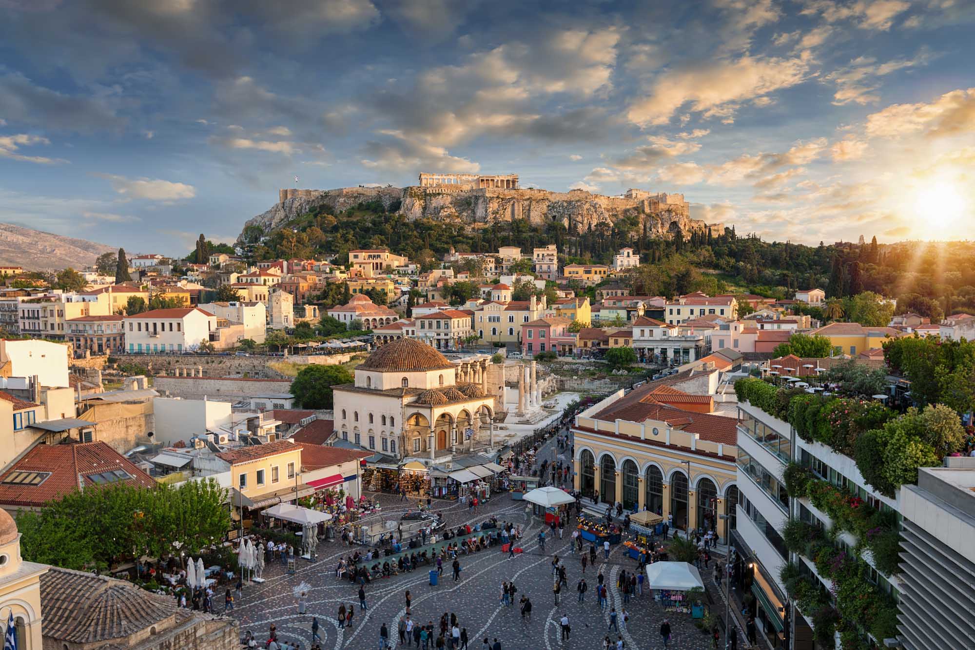 Sunset Over Plaka with Acropolis, Athens A stunning golden-hour view of Athens' historic Plaka district with colorful neoclassical buildings clustered below the illuminated Acropolis and Parthenon Temple perched atop a rocky hill. The scene captures the Byzantine Church of the Holy Apostles in the foreground with tourists gathered in the bustling square, exemplifying the blend of ancient history and vibrant modern life in Greece's capital.