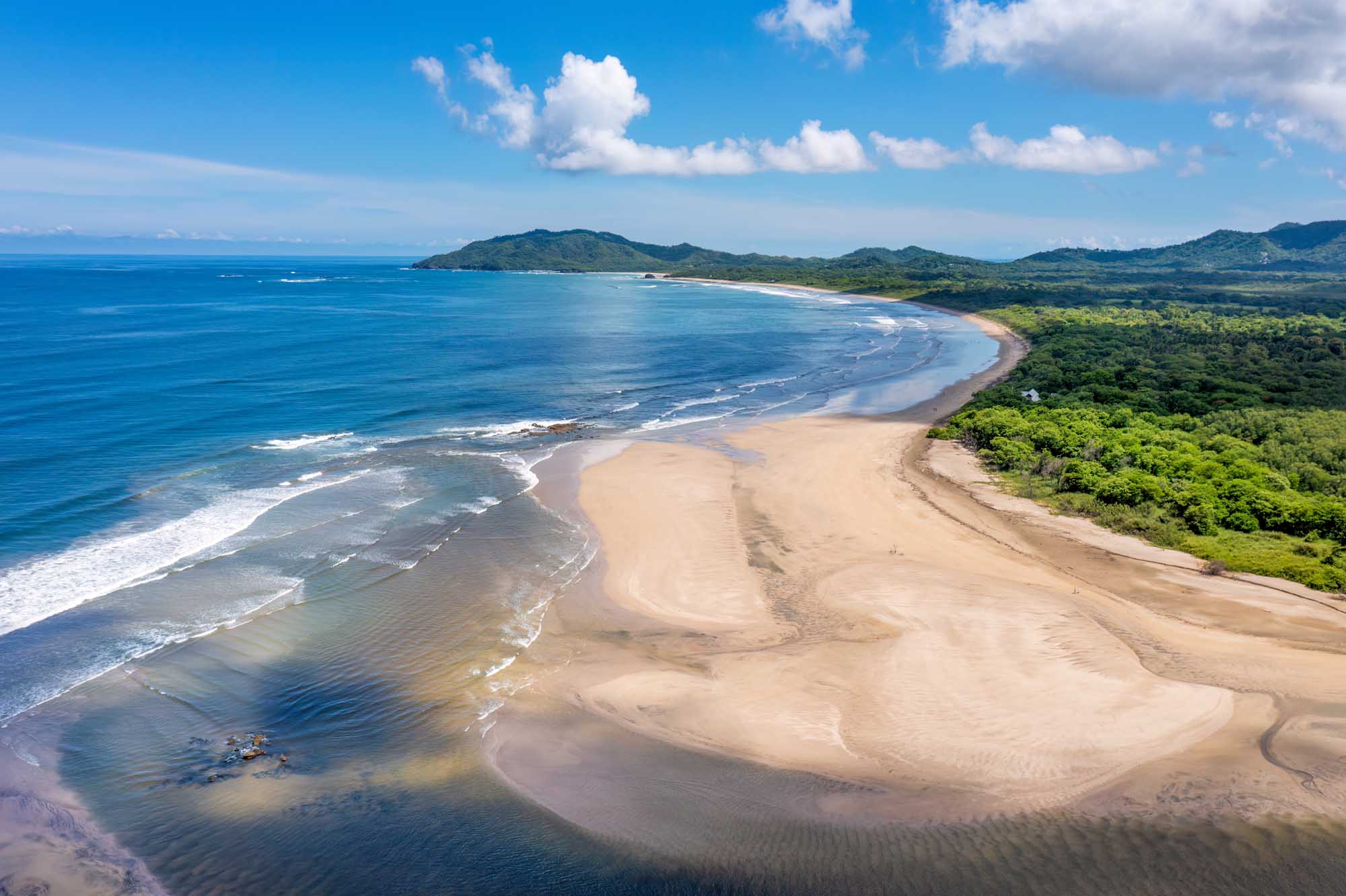 Tamarindo Beach and Estuary Aerial View An aerial view of Tamarindo Beach and the Tamarindo River estuary in Guanacaste, Costa Rica, showing the pristine sandy beach curving along the Nicoya Peninsula with lush tropical vegetation, river mouth, and turquoise Pacific waters with white-capped waves under a bright blue sky.