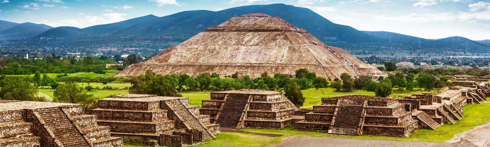 Teotihuacan Pyramids and Ancient Ruins The iconic Pyramid of the Sun dominates this expansive view of Teotihuacan's archaeological complex in Mexico, with smaller pyramidal structures and ancient foundations scattered across verdant grounds, set against blue mountains and a clear sky.