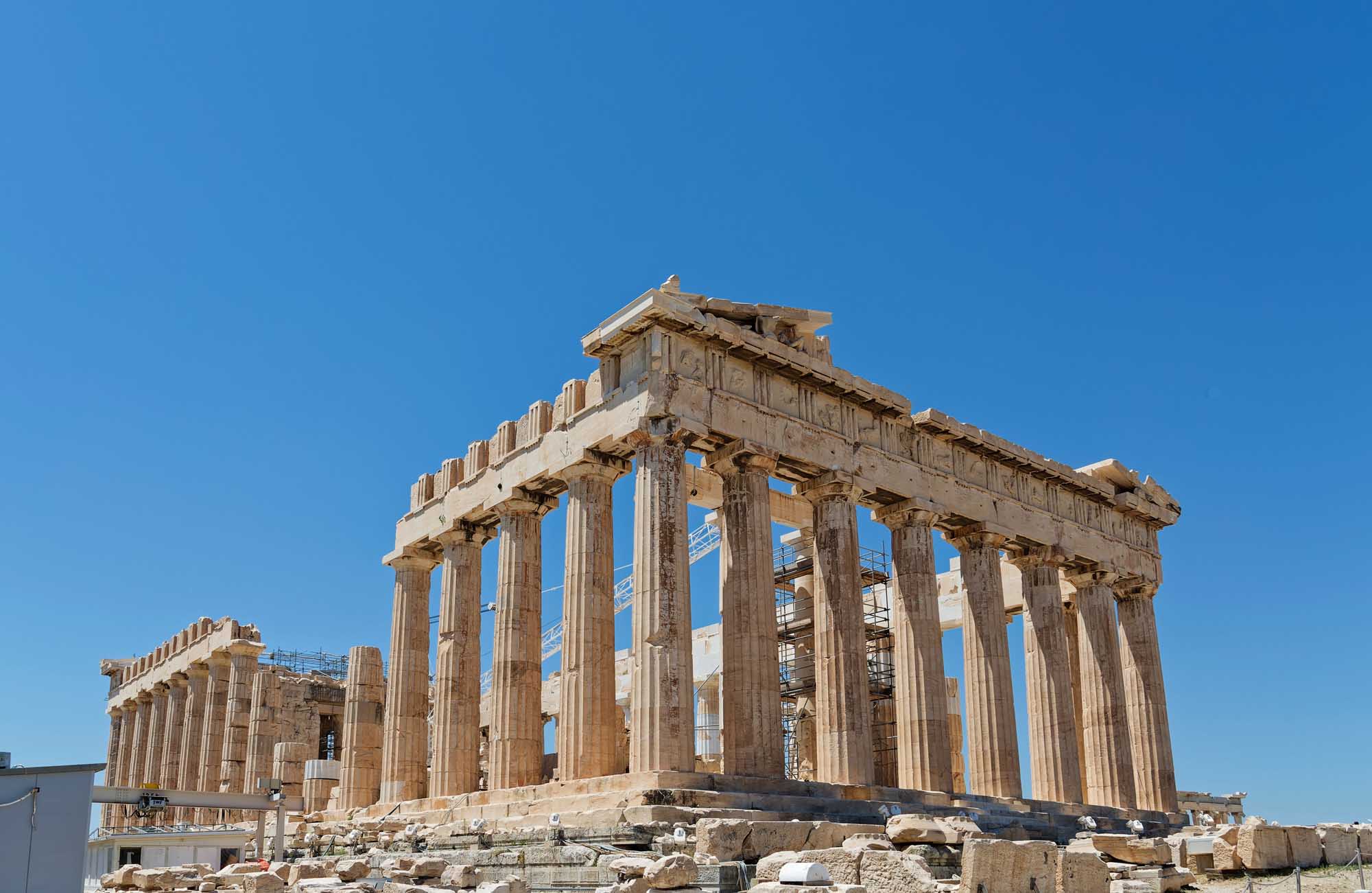 The Parthenon Temple on the Acropolis The iconic Parthenon temple stands majestically on the Athenian Acropolis against a clear blue sky. The ancient Greek structure features its distinctive Doric columns and ornate architectural details, with visible restoration scaffolding indicating ongoing preservation work.
