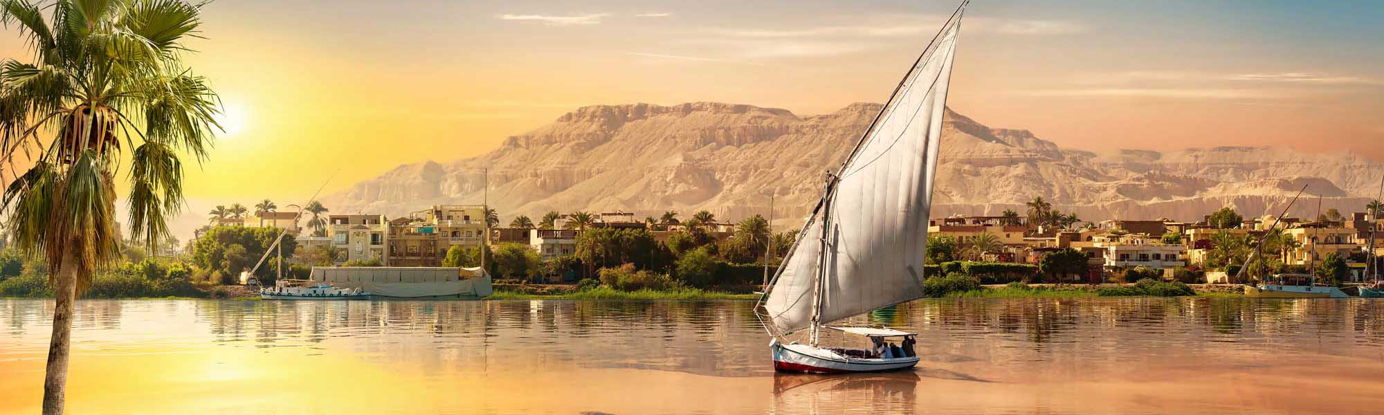 Traditional Felucca Sailing on the Nile River A traditional Egyptian felucca sailboat with white canvas sails glides peacefully across the Nile River at golden sunrise, with palm-lined riverbanks, colorful buildings, and desert cliffs in the background. The warm morning light bathes the scene in golden and amber tones, capturing the timeless beauty of river travel in Egypt.