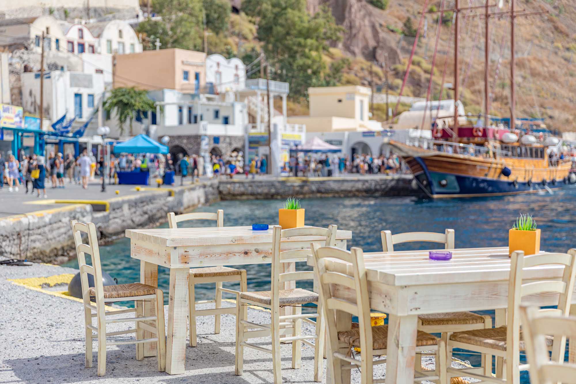 Traditional Greek Taverna at Santorini Harbor A charming waterfront taverna in Santorini, Greece, featuring whitewashed wooden tables and chairs with woven rush seats overlooking a bustling harbor. The scene captures traditional Cycladic architecture with blue-trimmed buildings, moored sailing vessels, and tourists exploring the picturesque port village.