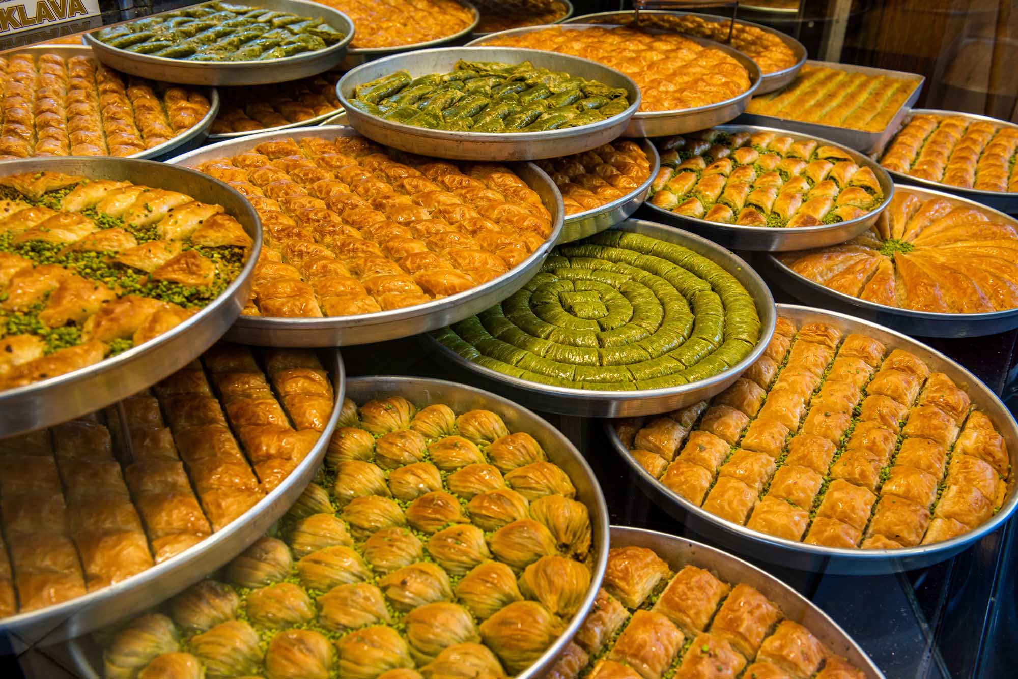 Traditional Turkish Baklava Display at Dessert Shop A vibrant display of various traditional Turkish baklava varieties arranged in metal trays at a dessert shop. The golden pastries are garnished with pistachios and arranged in decorative patterns, showcasing different baklava styles including spiral-shaped pistachio baklava and rectangular honey-soaked varieties typical of Gaziantep, Turkey.