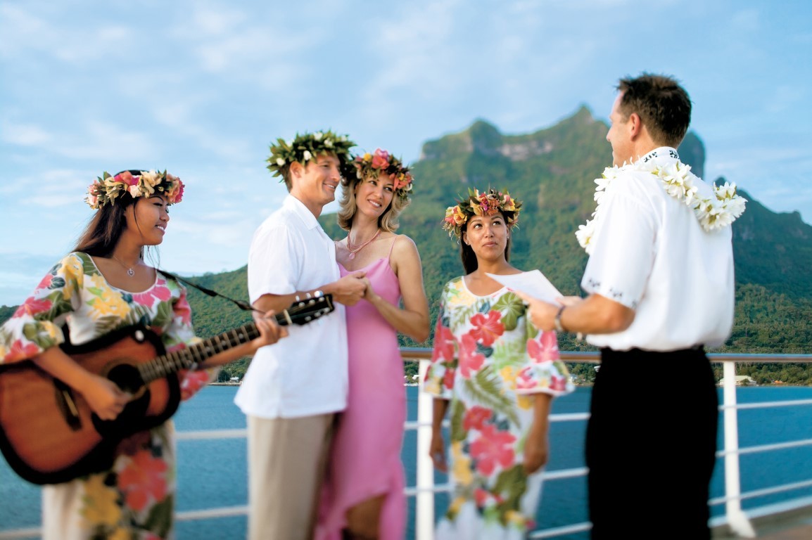 Tropical Island Wedding Celebration on Ship A joyful wedding celebration on a cruise ship's deck featuring five people in tropical attire and flower garlands, with a lush green volcanic island and turquoise ocean visible in the background. The scene captures a Hawaiian or South Pacific-themed destination wedding moment.