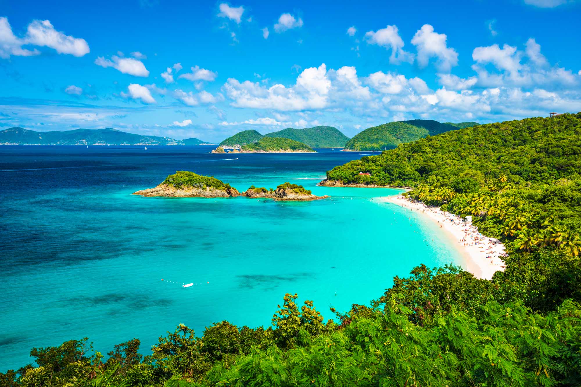 Trunk Bay Beach, St. John, US Virgin Islands A breathtaking tropical paradise featuring pristine turquoise waters, white sandy beaches, and lush green vegetation. Small rocky islands dot the crystalline bay while sailboats anchor in the calm water, with verdant hillsides and distant islands framing the spectacular seascape under a brilliant blue sky.