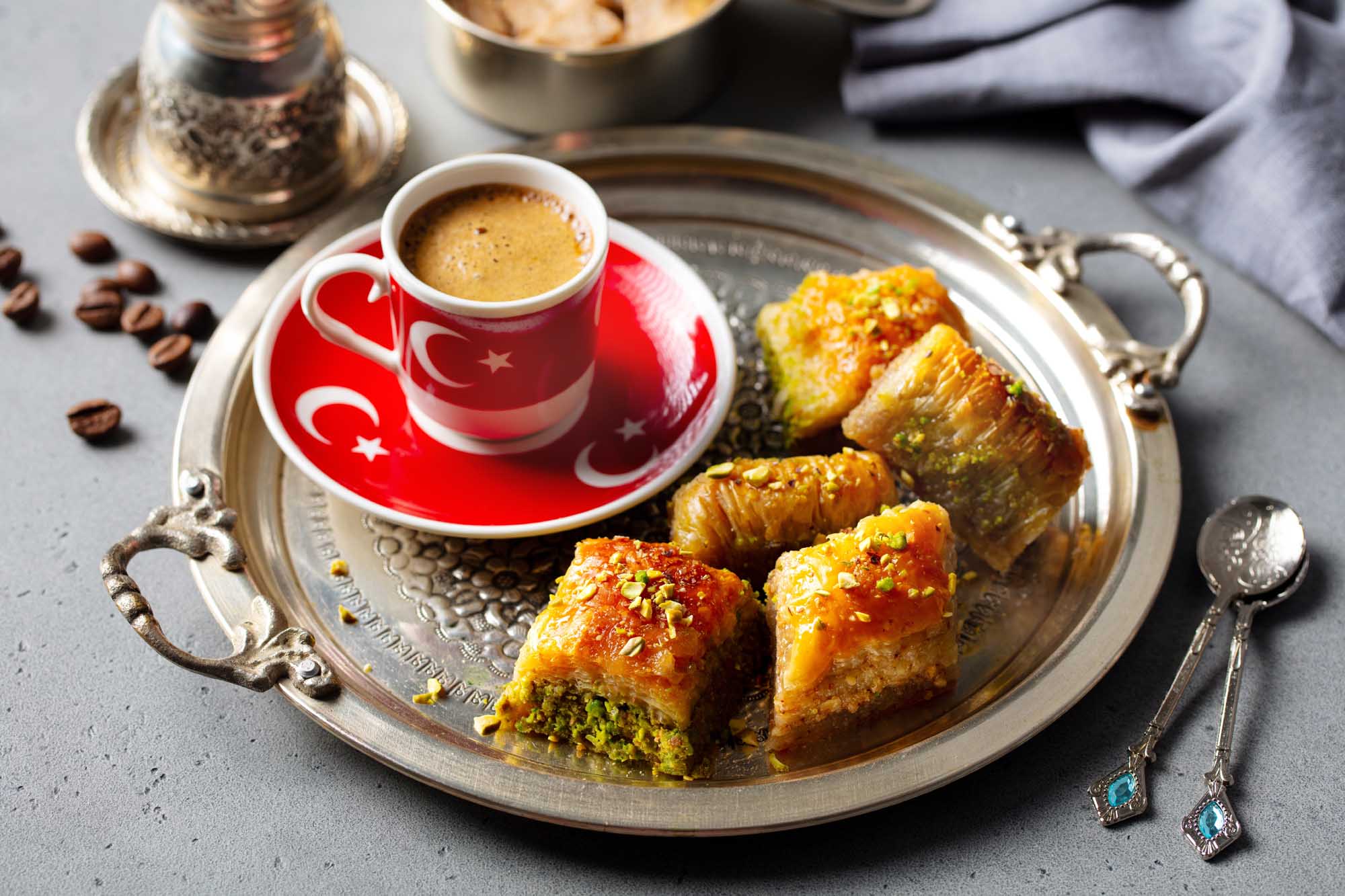 Turkish Coffee with Baklava Pastries An elegant presentation of traditional Turkish coffee served in a red cup with Turkish flag motifs, accompanied by golden baklava pastries topped with pistachios on an ornate silver tray. The composition includes scattered coffee beans, decorative silver utensils, and traditional Turkish coffee serving accessories, showcasing authentic Middle Eastern culinary culture.