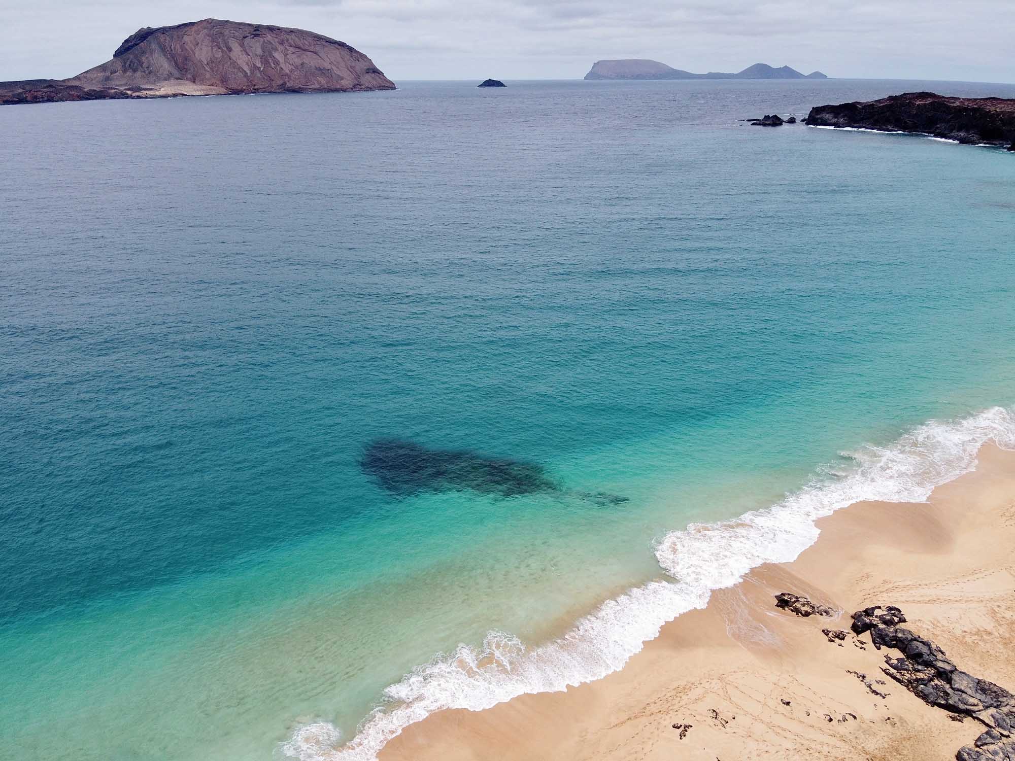 Turquoise Beach with Volcanic Islands A pristine sandy beach with crystal-clear turquoise waters and gentle white-capped waves, featuring dramatic volcanic islands visible in the distance across a calm sea. The landscape showcases volcanic rock formations along the shoreline and a distinctive cone-shaped island on the left horizon.