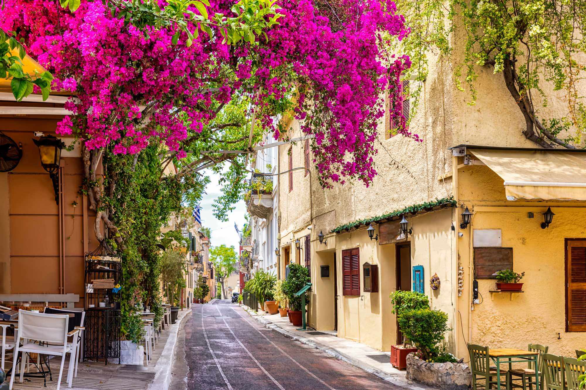 Vibrant Mediterranean Street with Bougainvillea A charming Mediterranean alleyway lined with traditional stone buildings and cascading magenta bougainvillea flowers overhead, featuring characteristic architecture with colorful doors, shutters, and outdoor café seating along a quiet street.